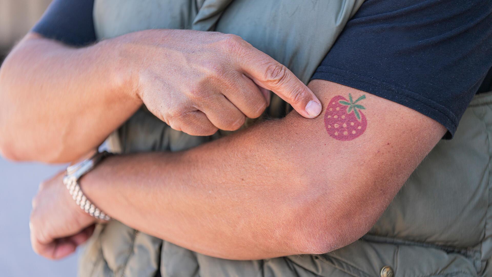 Ein Mann zeigt auf ein Erdbeer-Tattoo auf seinem linken Arm.