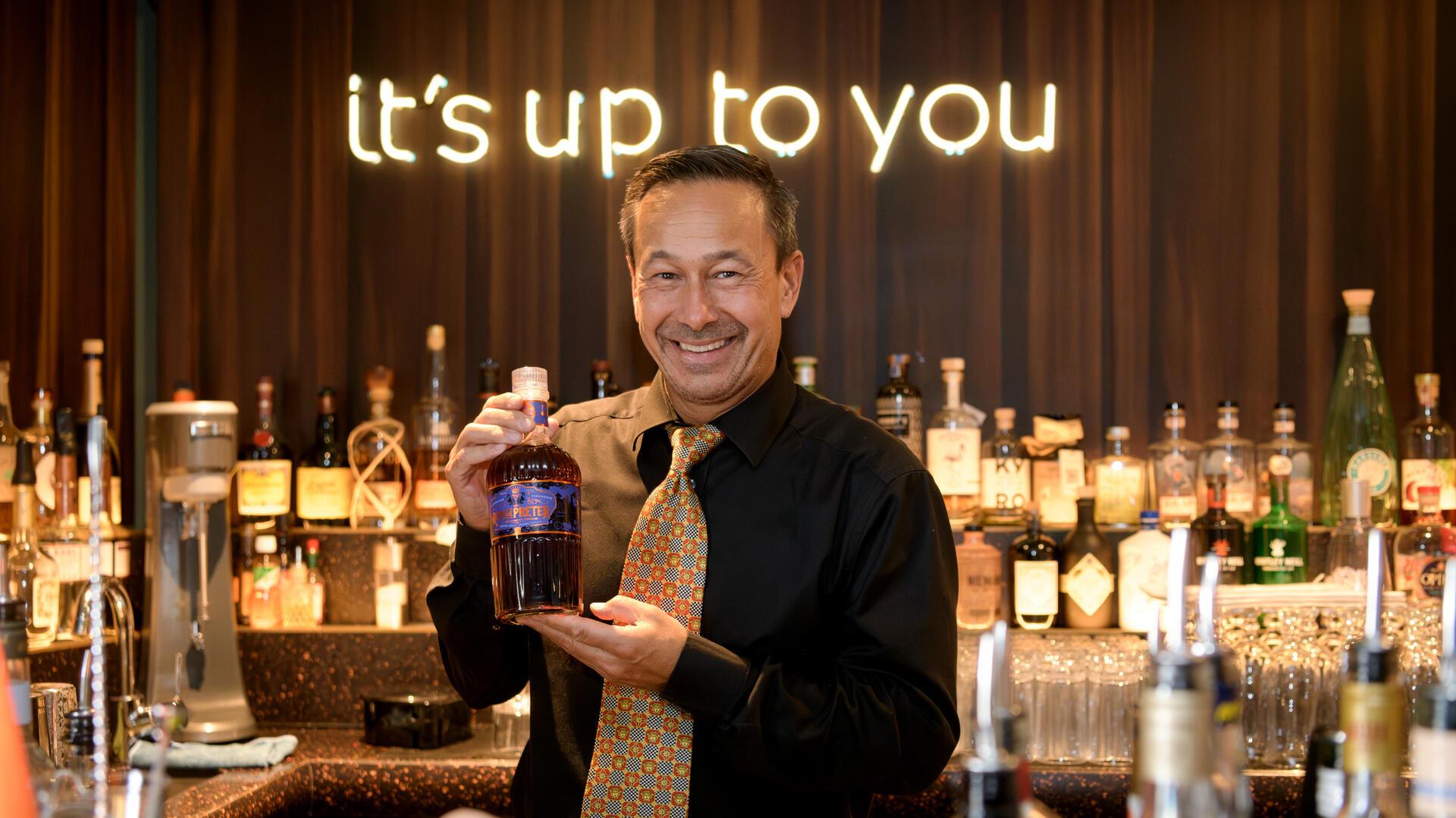 Ein Mann in einer Bar, der seine Rumkreation zeigt. Im Hintergrund ist in leuchtender Schrift "it's up to you" zu lesen. 