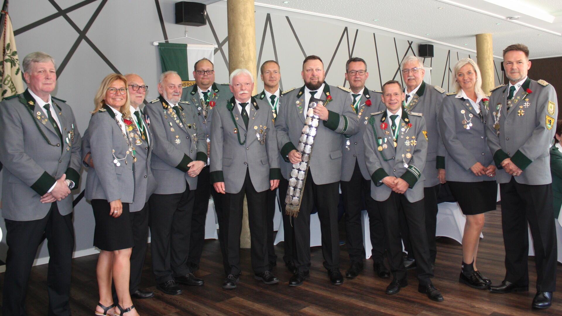 Gruppenfoto von Mitgliedern eines Schützenvereins in festlicher Uniform mit Orden und Abzeichen. In der Mitte steht der Schützenkönig mit der Schärpe. Die Personen posieren in einem Saal mit Schützenfahne und dekorativer Wand im Hintergrund.