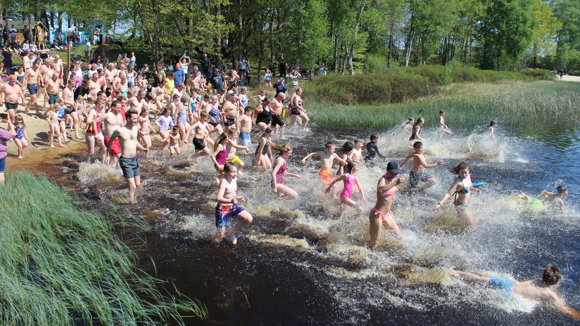 Zahlreiche Menschen, darunter viele Kinder, rennen in Badekleidung bei sonnigem Wetter gemeinsam ins Wasser eines Sees. Am Ufer stehen Zuschauer, im Hintergrund sind Bäume und ein Sandstrand zu sehen.