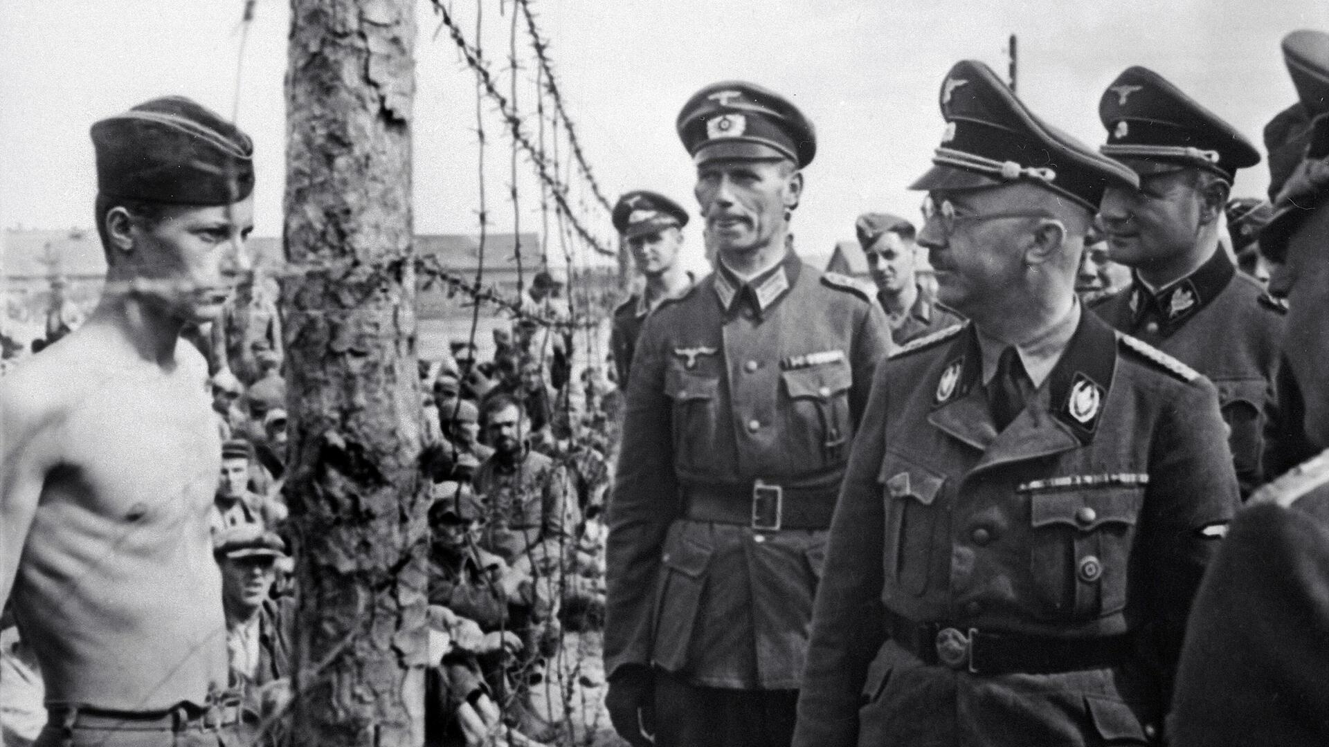 SS-Chef Heinrich Himmler blickt einen Kriegsgefangenen hinter einem Zaun an.