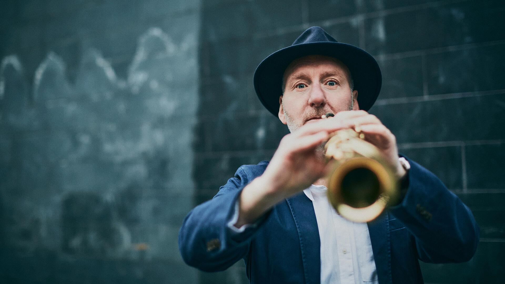 Ein Mann mit Hut spielt Saxophon und blickt direkt in die Kamera. Die Aufnahme ist leicht unscharf im Vordergrund und zeigt eine dunkle Wand im Hintergrund.