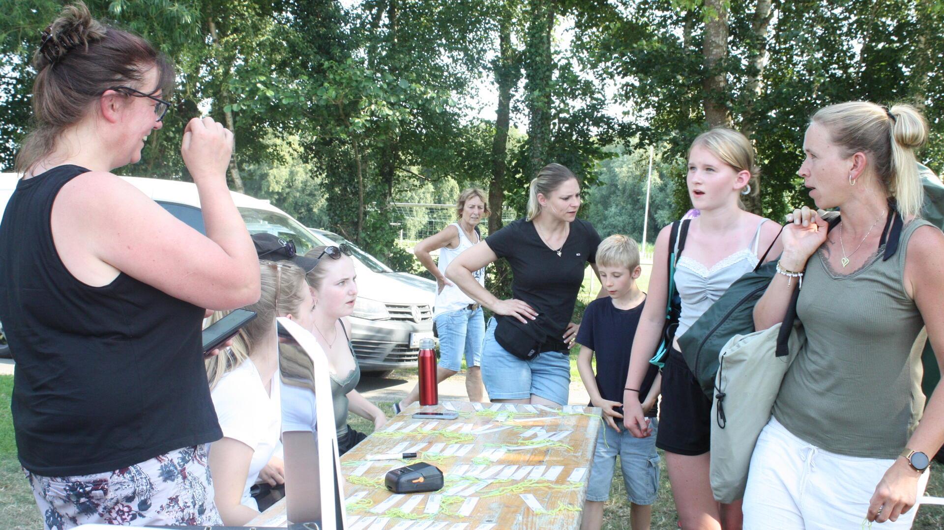 Eltern und Kinder stehen an einem Anmeldetisch im Freien, umgeben von Bäumen. Auf dem Tisch liegen Namensschilder, im Hintergrund sind Erwachsene und Fahrzeuge zu sehen.