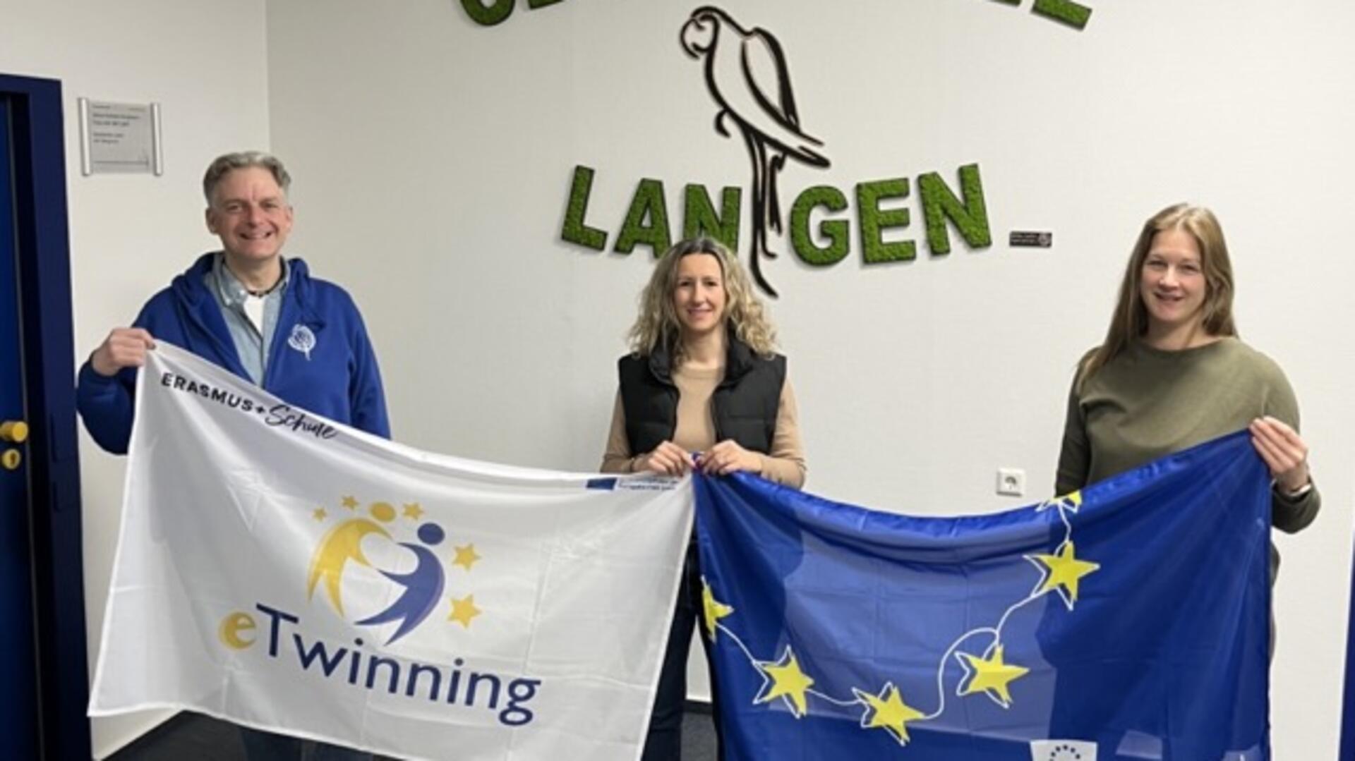 Ein Mann und zwei Frauen stehen nebeneinander. Sie halten gemeinsam zwei Flaggen mit ihren Händen.