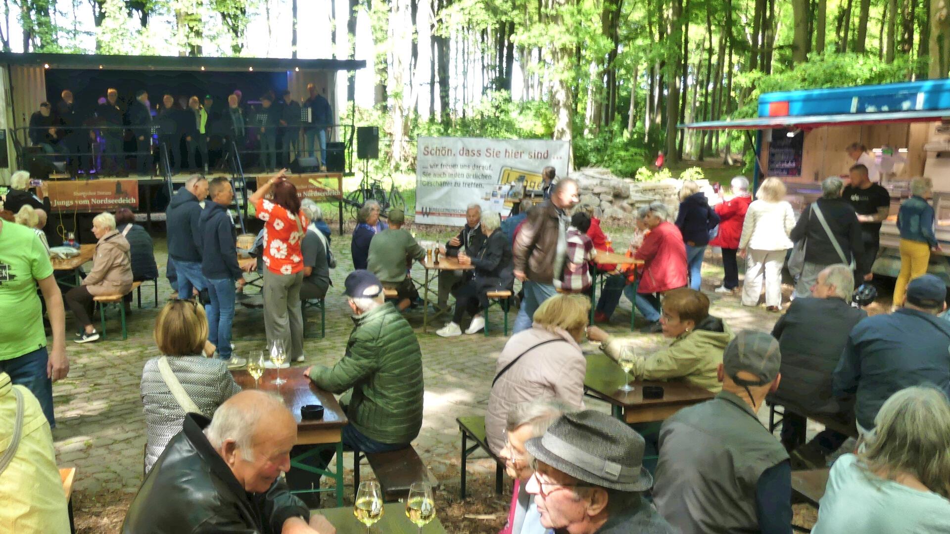 Menschen sitzen und stehen bei sonnigem Wetter im Park Friedrichsruh auf Bierbänken vor einer Bühne, auf der der Shantychor „Jungs vom Nordseedeich“ auftritt; im Hintergrund ein Imbisswagen und Waldkulisse.