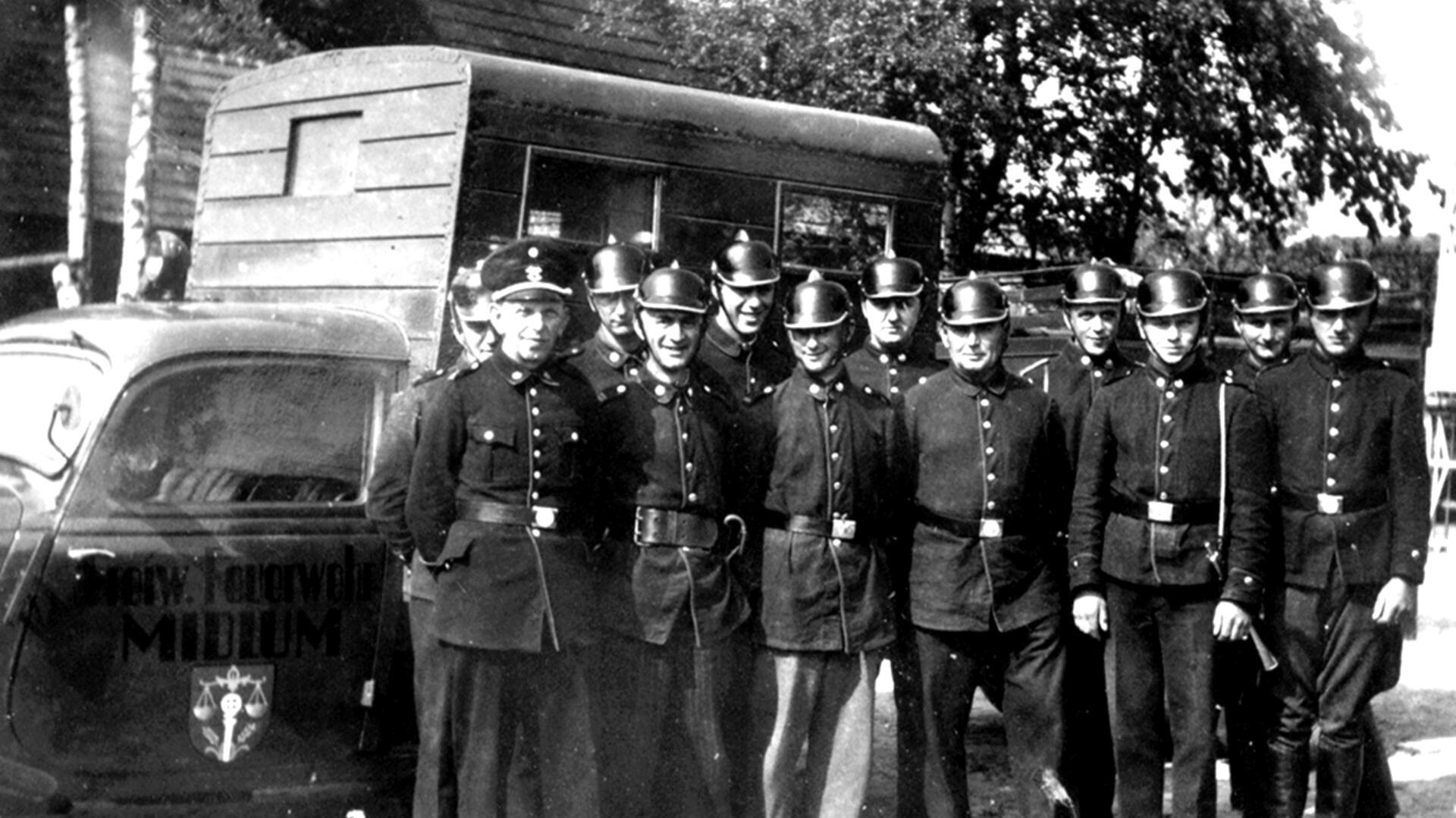 Schwarzweißfoto aus der Mitte des 20. Jahrhunderts: Eine Gruppe von Feuerwehrmännern der Freiwilligen Feuerwehr Midlum steht in Uniform und mit Helmen vor einem Einsatzfahrzeug.