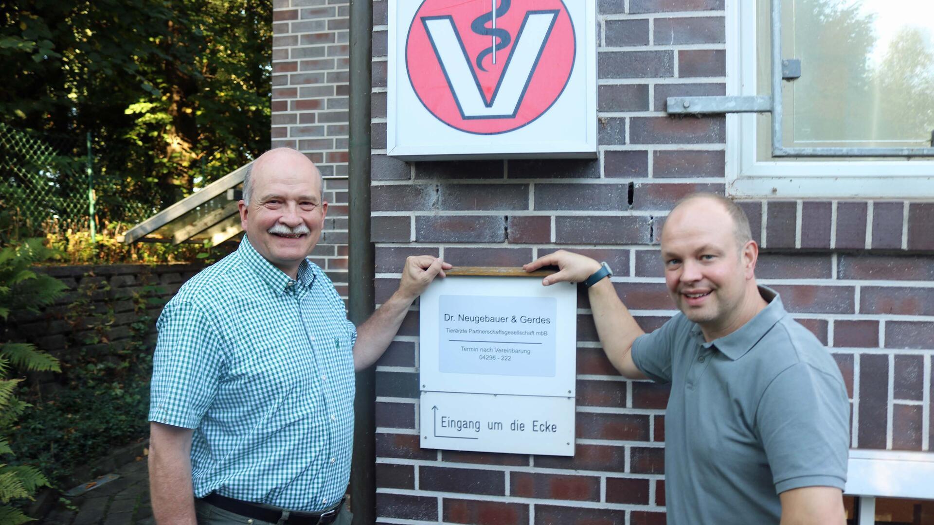Zwei Männer stehen vor einem Praxisschild