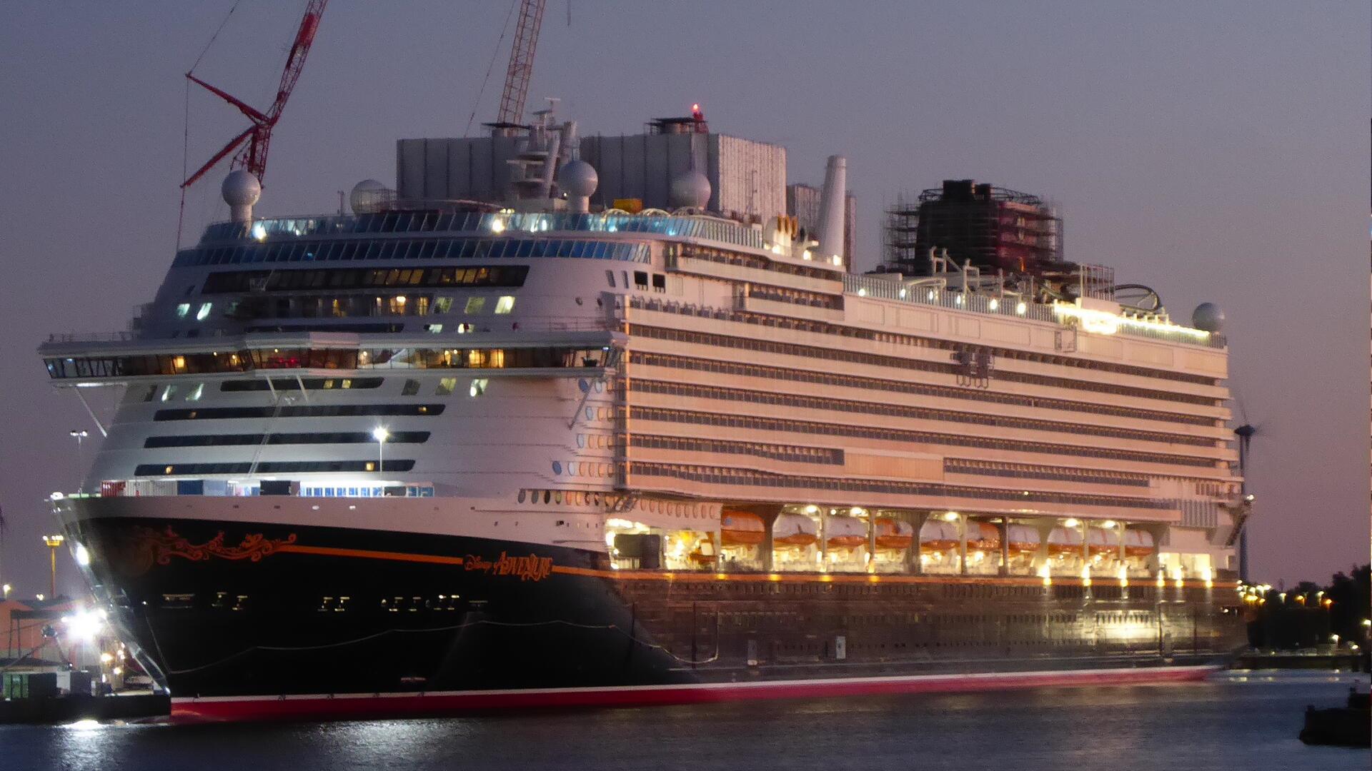 Die „Disney Adventure“ liegt beleuchtet am Abend im Bremerhavener Überseehafen an der Lloyd Werft. Auf dem Deck sind Baugerüste und Kräne zu erkennen.