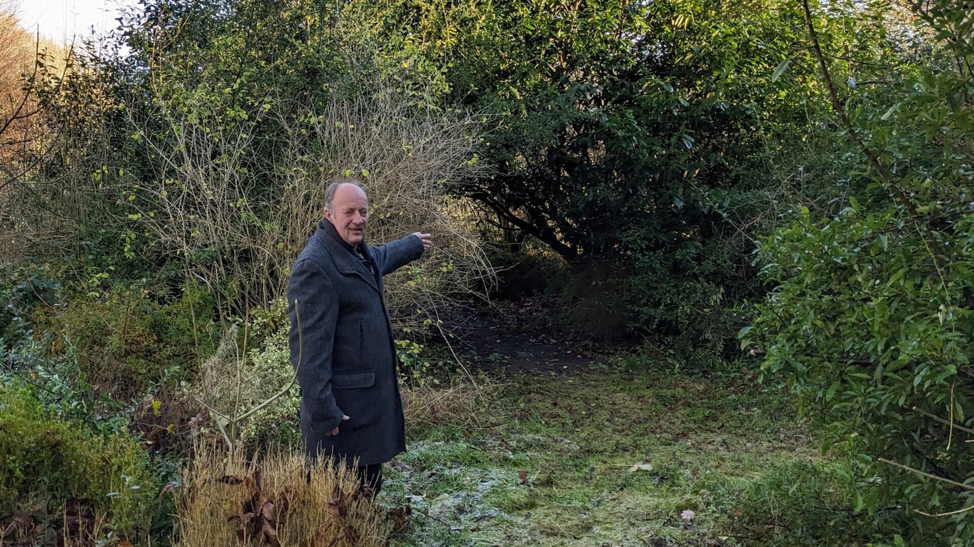 Ein Mann im dunklen Mantel steht in einem überwucherten Gartenbereich und zeigt auf ein zugewachsenes, dichtes Gebüsch.