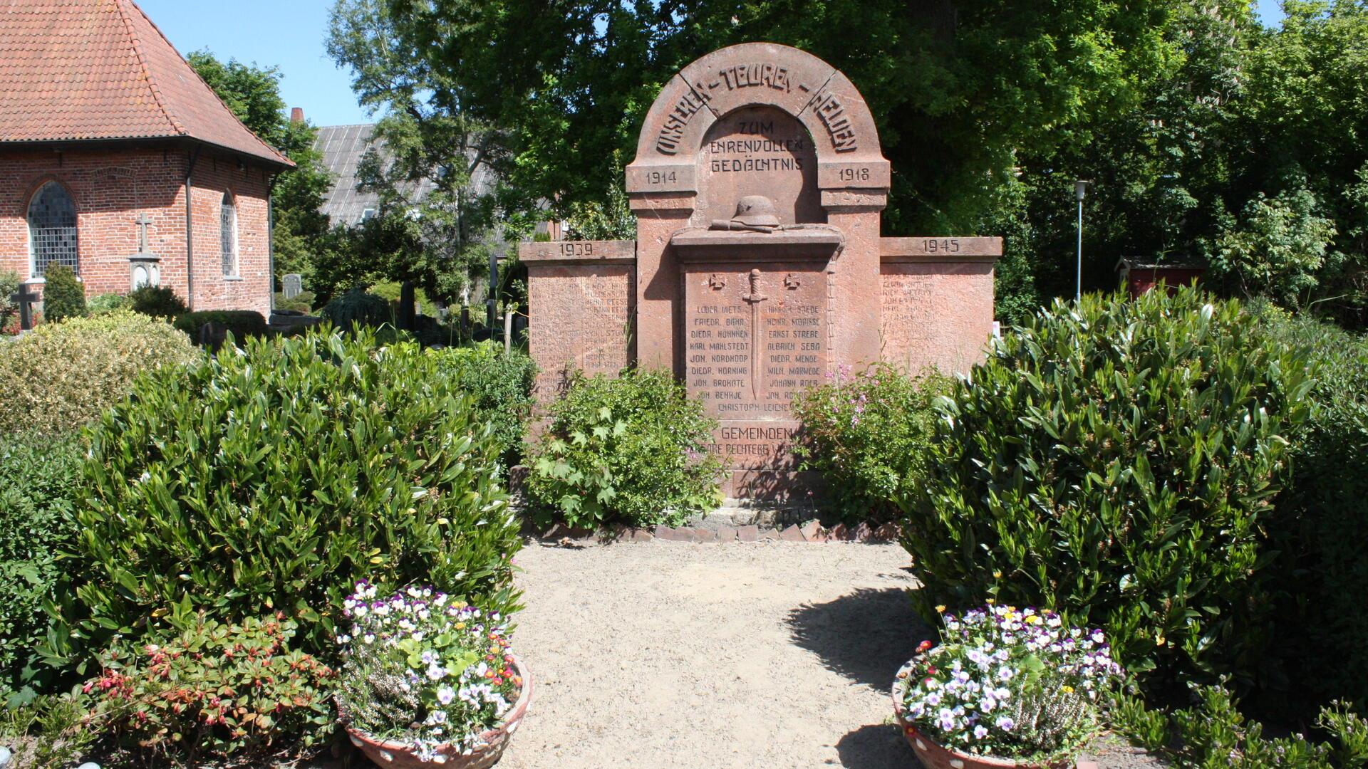 Ein Kriegerdenkmal im HintergrundDrumherum Sträucher und Blumenkübel.