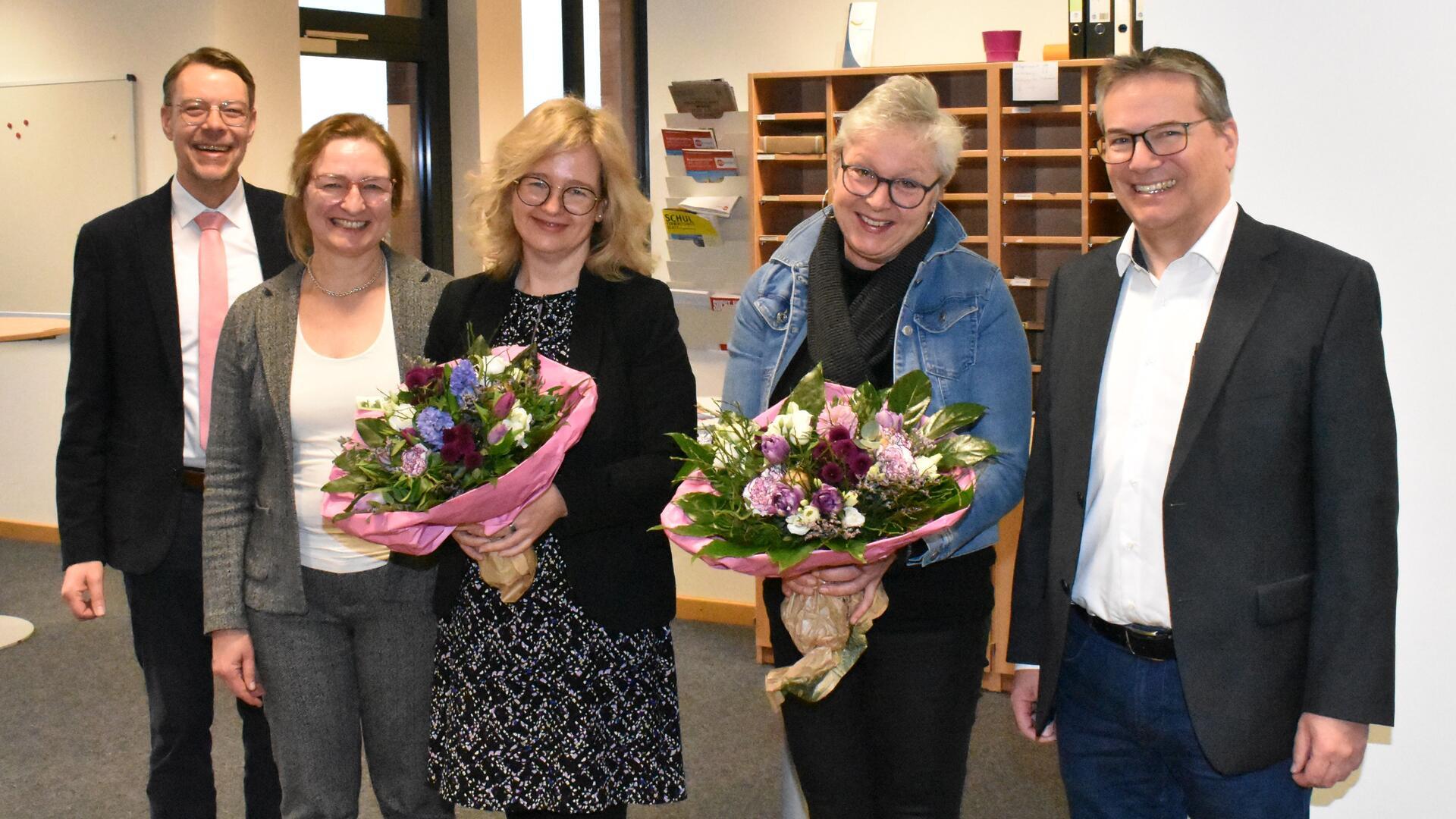 Fünf Personen nebeneinander. Links ein Mann, rechts daneben drei Frauen, zwei halten einen Blumenstrauß in den Händen und ganz rechts steht ein Mann.