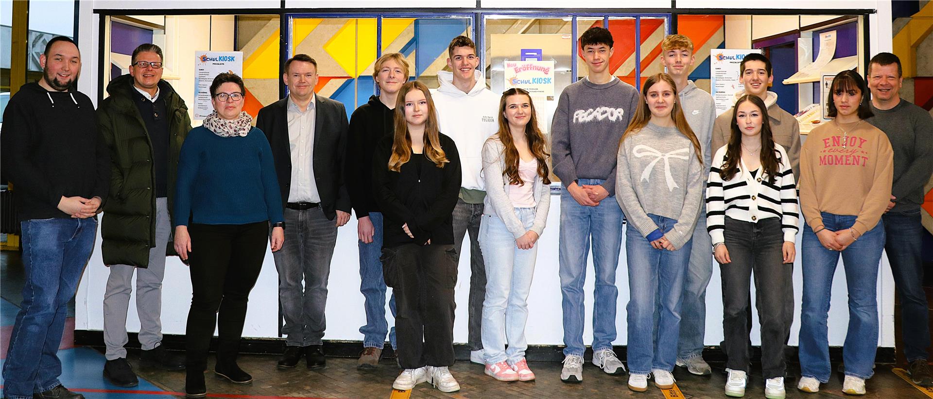 Eine Gruppe aus Jugendlichen und Erwachsenen steht vor einem Schul-Kiosk mit bunten Fenstern. Einige tragen lässige Kleidung, andere formelle Outfits. Im Hintergrund hängen Schilder zur Neueröffnung des Kiosks.