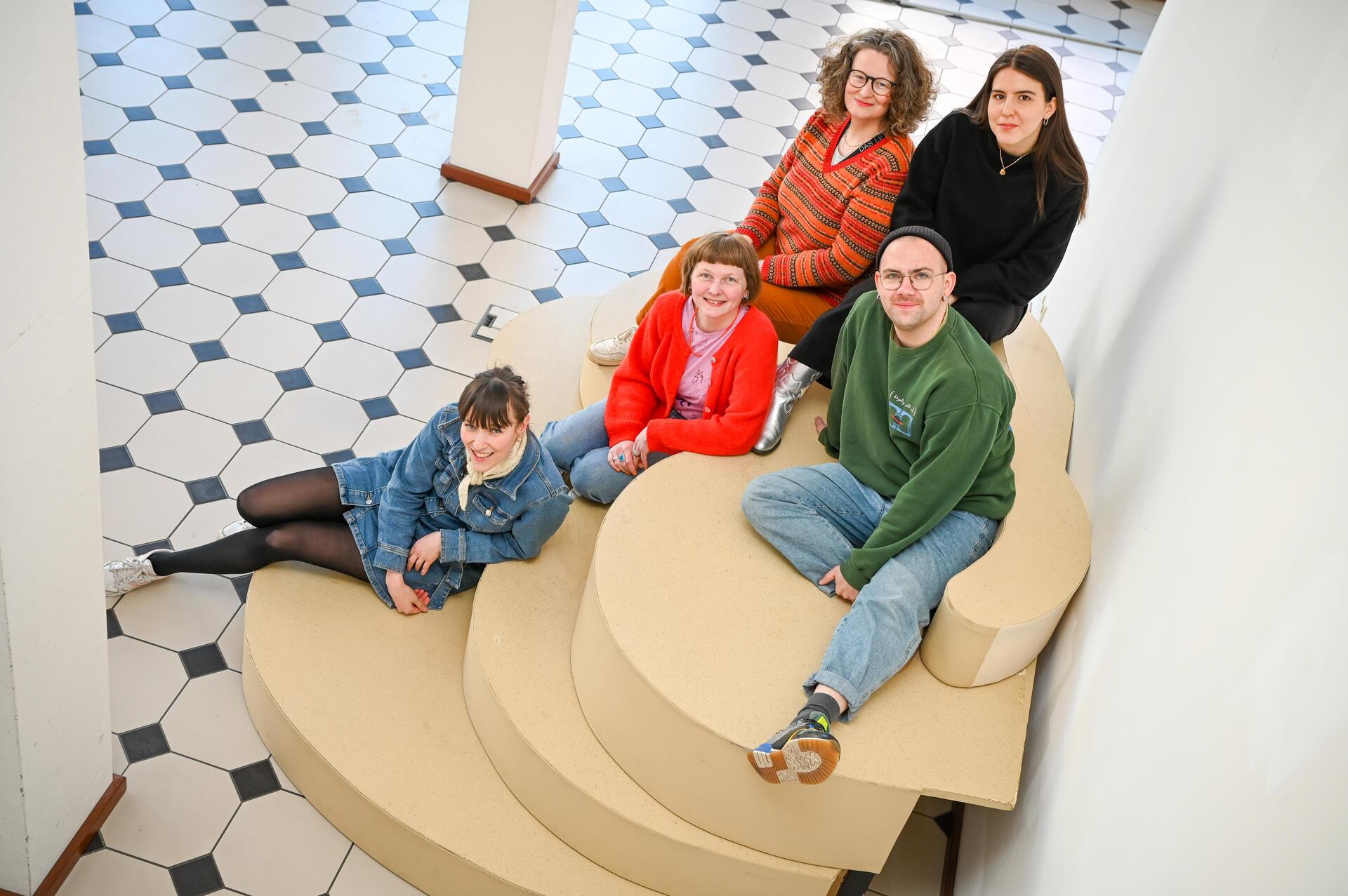 Fünf Menschen sitzen auf einer treppenähnlichen Couch. Sie lächeln und freuen sich nicht nur über die neuen Räume in der Fährstraße, sondern auch auf die anstehenden Premieren der Theaterlabore (von links): die Theaterpädagoginnen Katharina Dürr und Julia Balzert, JUB-Leiterin Bianca Sue Henne, Theaterpädagoge Steven Wind und seine Kollegin Schirin Badafaras. 