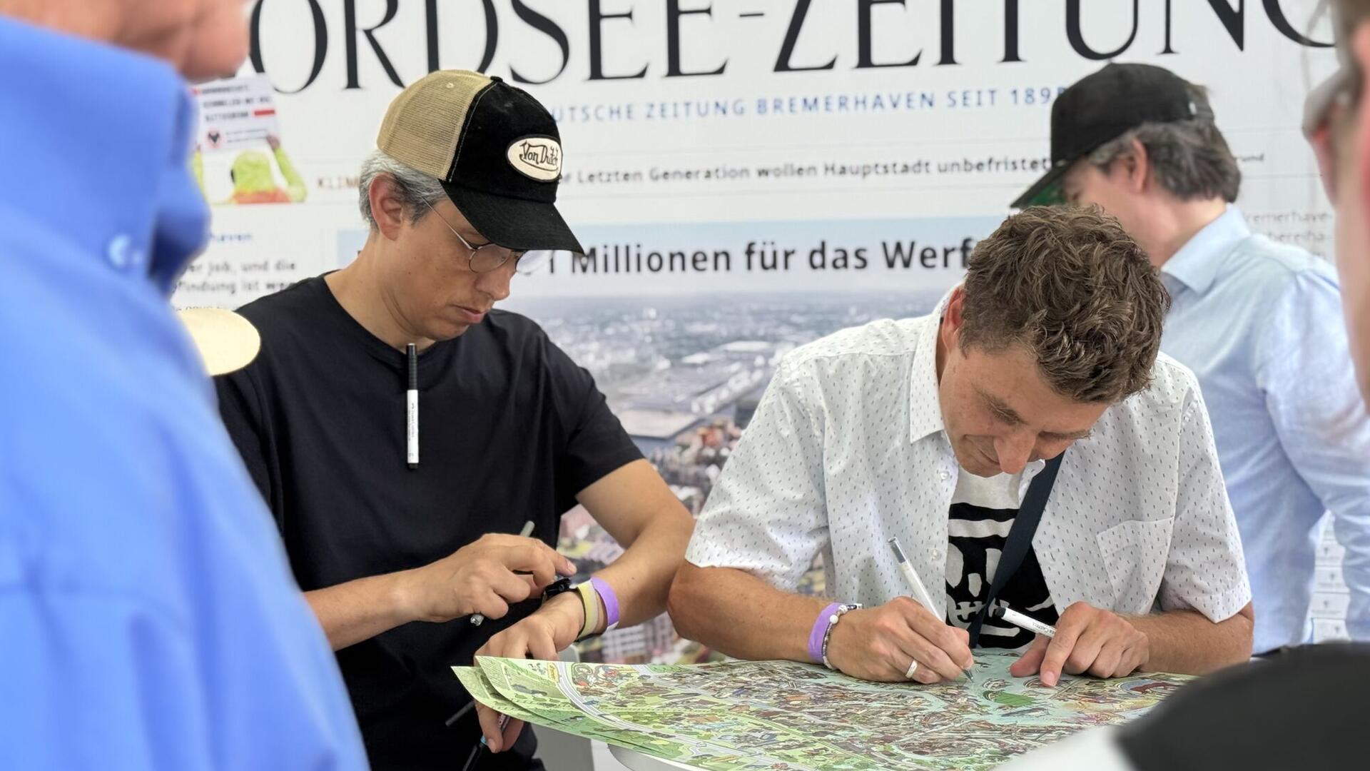 Zwei Männer stehen vor einem Poster, das auf einem hohen Tisch liegt. Einer von ihnen zeichnet darauf.