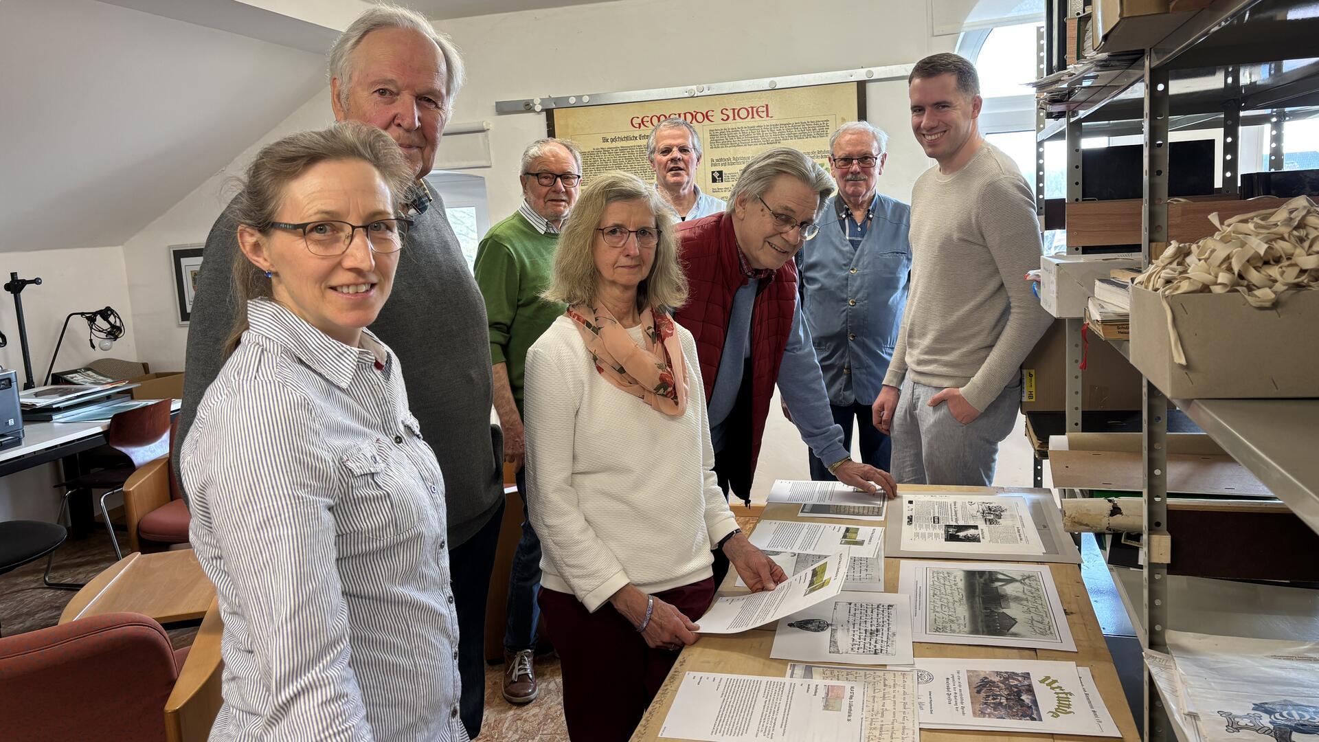 Eine Gruppe von acht Personen steht in einem Archivraum um einen Tisch mit historischen Dokumenten. Sie blicken in die Kamera, einige lächeln. Im Hintergrund sind Regale mit Akten und ein großes Informationsschild zur Geschichte von Stotel zu sehen.