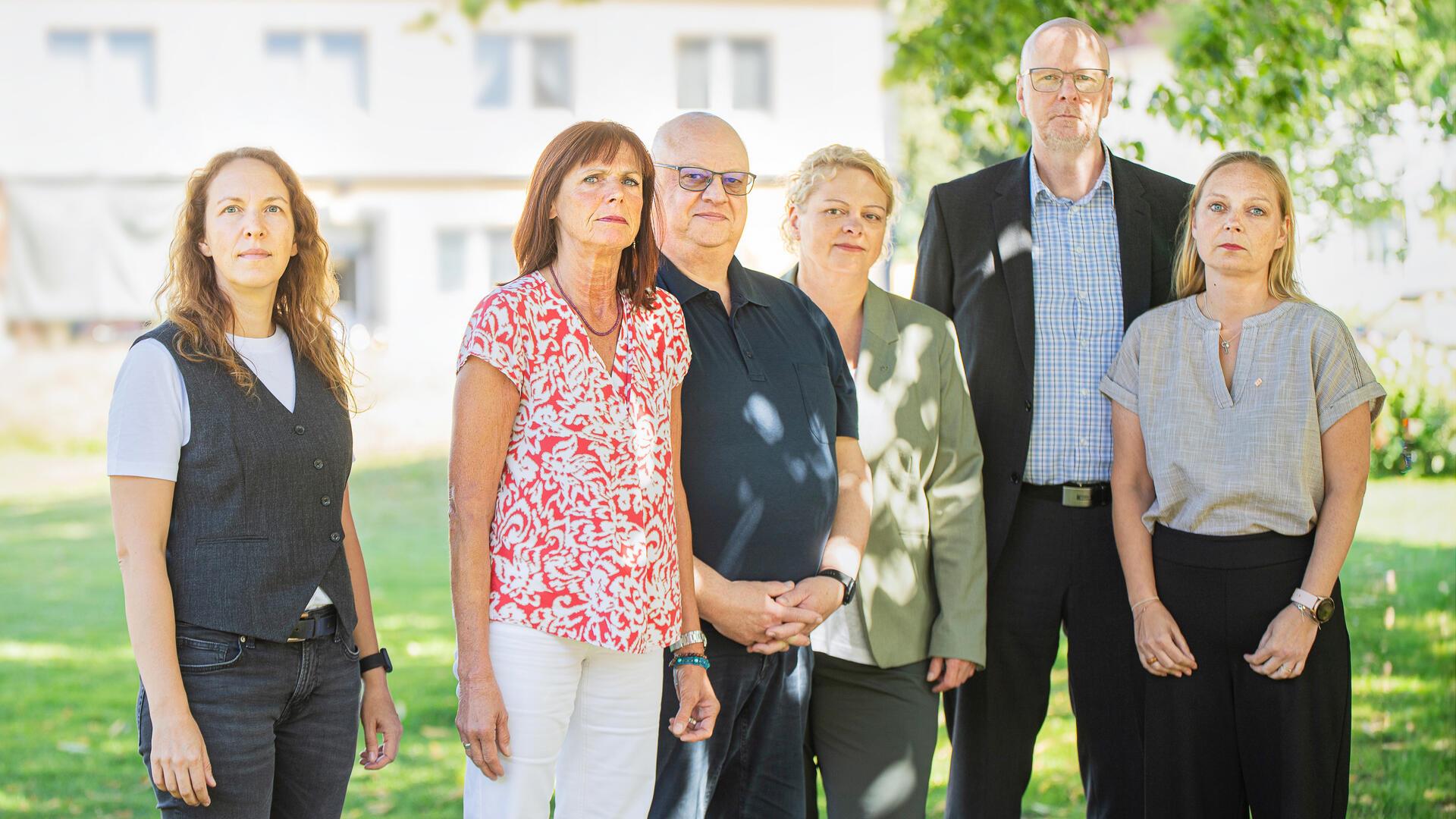 Sechs Personen stehen auf einer grünen Wiese unter einem Baum