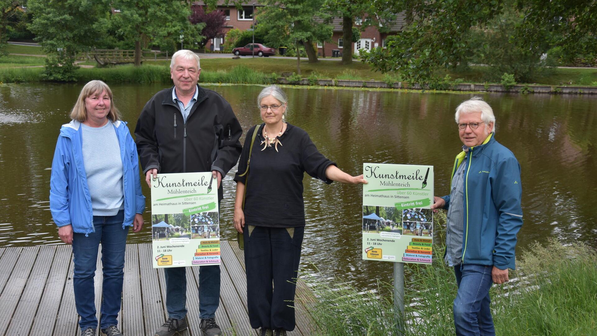 Zwei Frauen und zwei Männer stehen auf einem Steg an einem Teich und halten Plakate hoch.