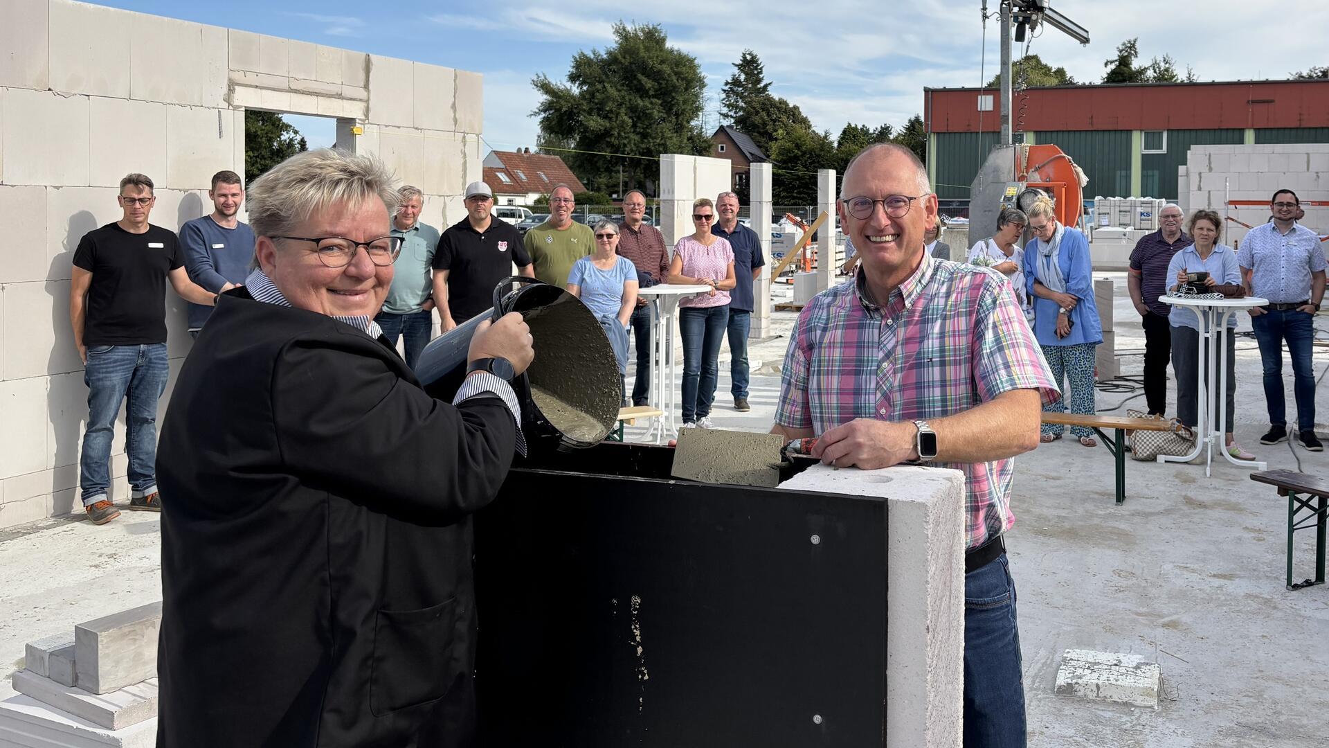 Eine Frau kippt einen Eimer mit Beton in eine Vorrichtung. Gegenüber steht ein lächelnder Mann in Hemd mit einer Kelle voller Beton.