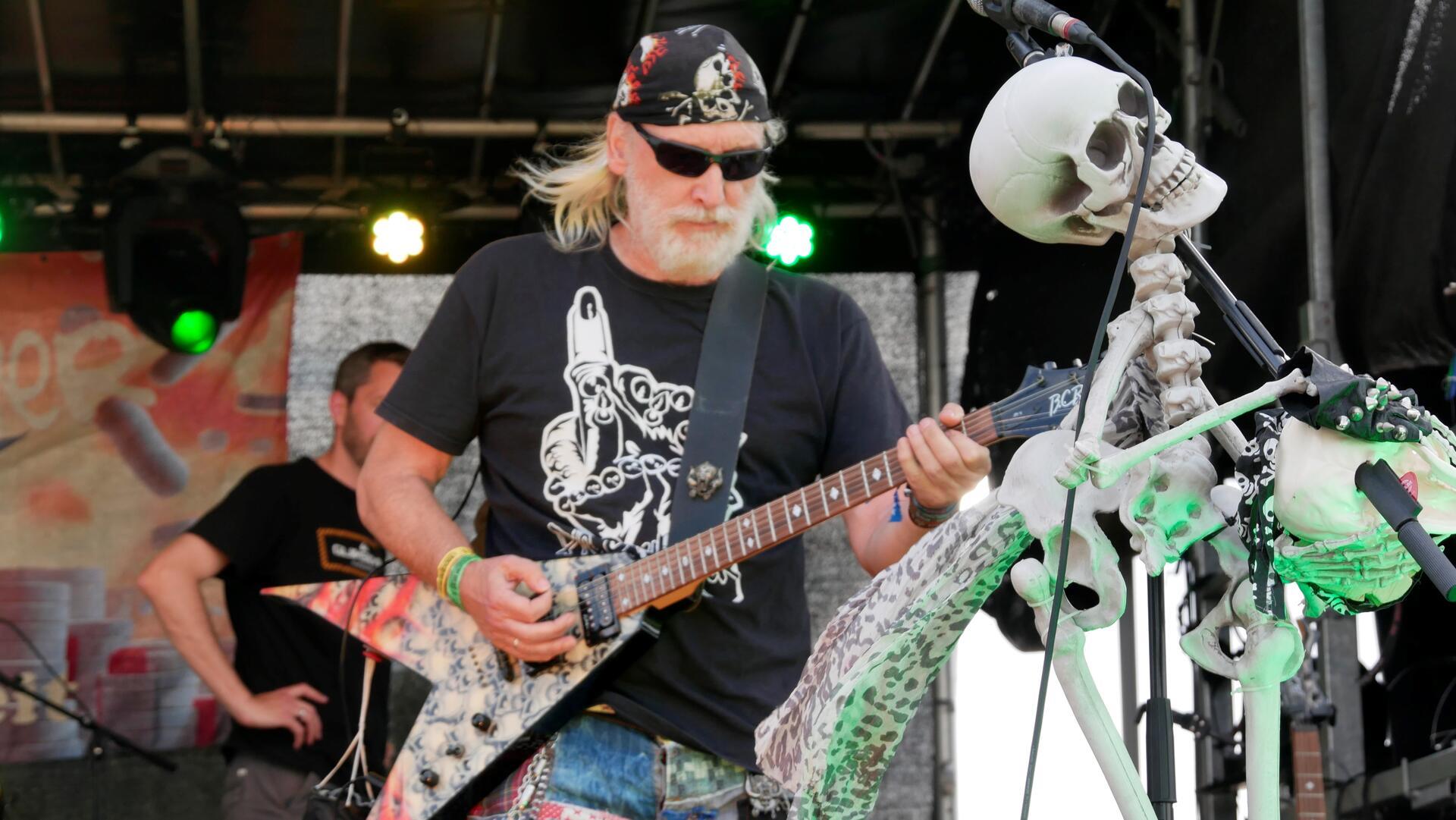 Ein Musiker hält eine Gitarre. Neben ihm steht ein künstliches Skelett. 