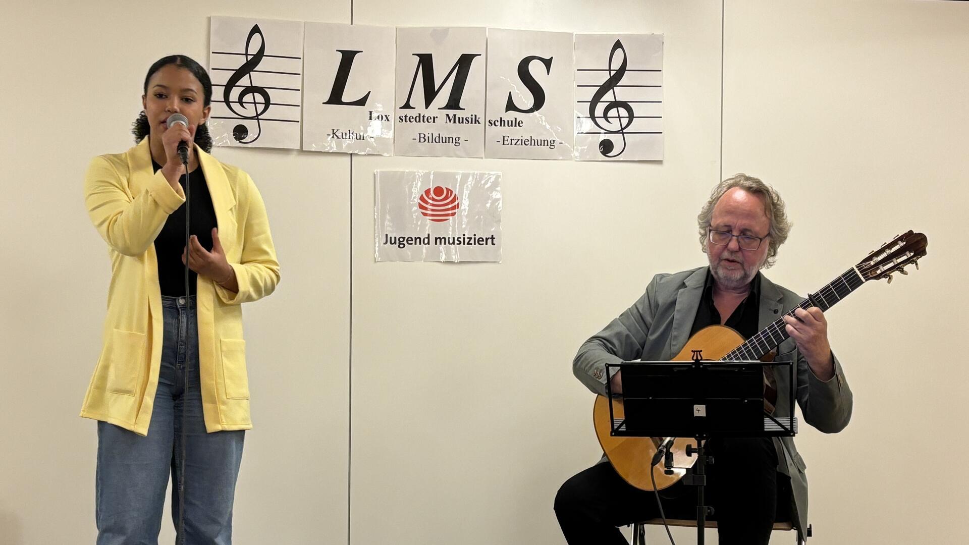 Eine junges Mädchen singt in ein Mikrofon, während ein älterer Mann sie auf einer klassischen Gitarre begleitet. Im Hintergrund hängen Schilder mit der Aufschrift „LMS – Loxstedter Musikschule“ sowie das Logo von „Jugend musiziert“.