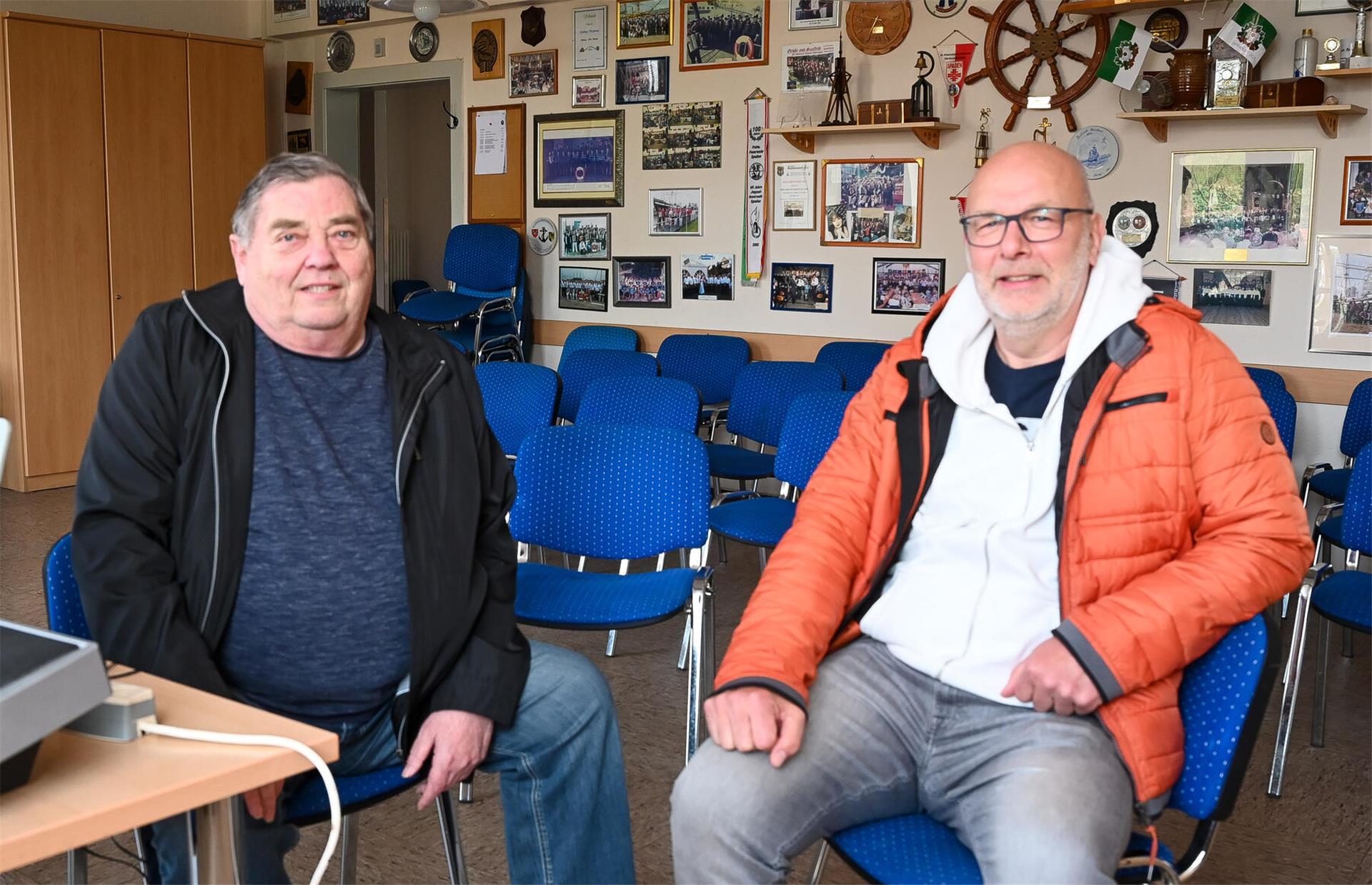 Zwei Männer, die jeweils auf Stühlen sitzen. Sie befinden sich in einem Raum, der mit Fotos und einem Schiffsrad an der Wand aufwartet.