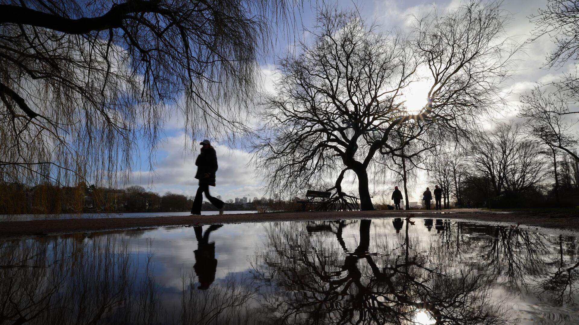 Bilder des Tages Spiegelung: Spaziergänger genießen den sonnigen Tag an der Alster in Hamburg, ihre Silhouetten und die der kahlen Bäume zeichnen sich in einer Pfütze ab.