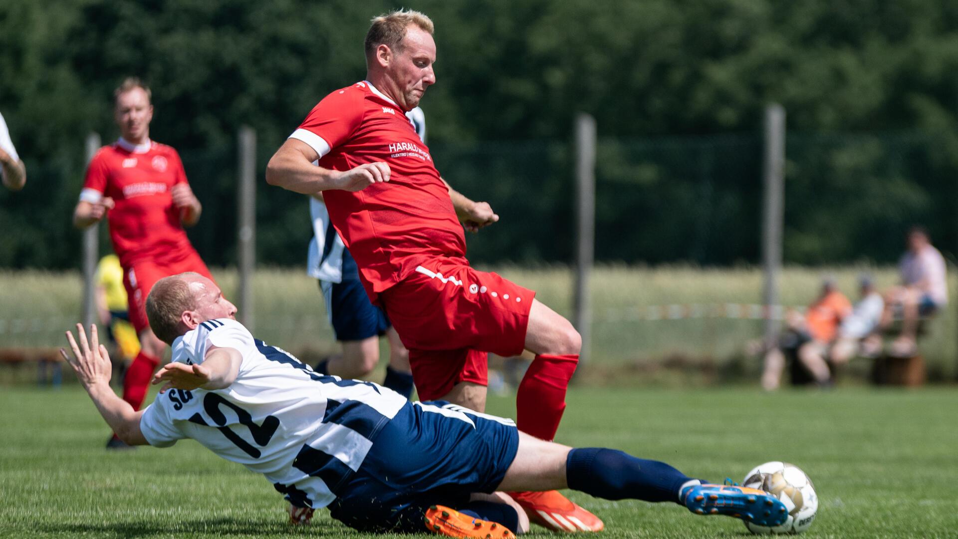 Zwei Männer in Sporttrikots versuchen einen Ball mit dem Fuß zu bekommen.
