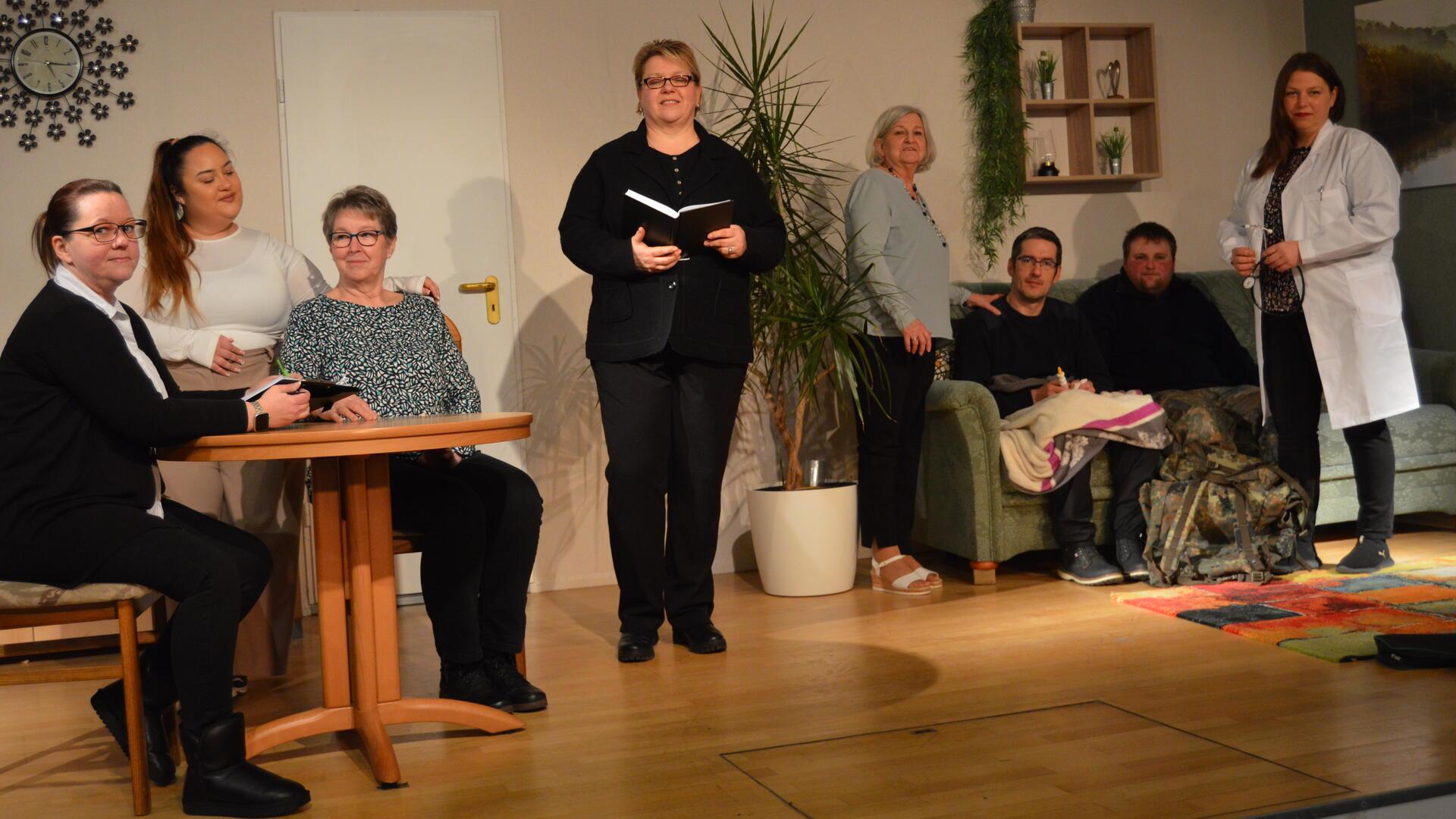 Eine Theatergruppe steht auf einer Bühne in der Kulisse eines Wohnzimmers. Einige Darsteller sitzen an einem Tisch, andere auf einem Sofa, während eine Person in einem weißen Arztkittel steht. 