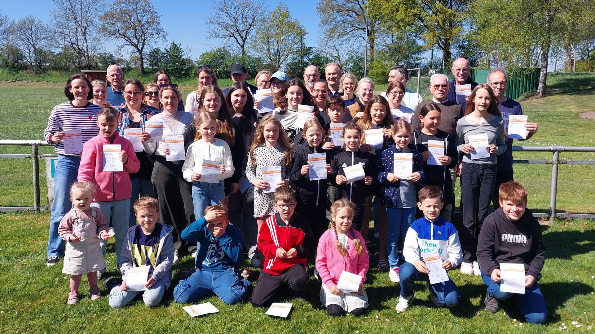 Gruppenfoto vor einem Sportplatz: etwa 30 Teilnehmende aller Altersstufen von Kleinkindern bis Senioren stehen bei sonnigem Wetter auf einer Wiese und halten stolz ihre Urkunden für das Deutsche Sportabzeichen in den Händen.