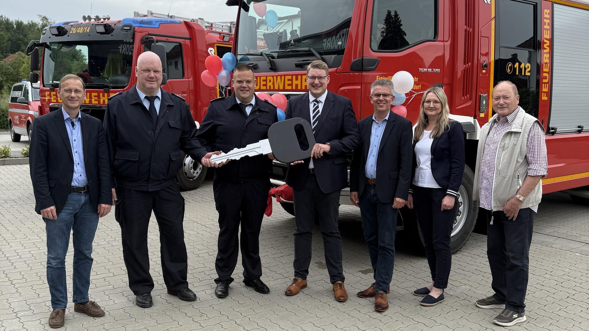 Vertreter von Feuerwehr, Gemeinde und Politik stehen vor einem neuen Feuerwehrfahrzeug. Zwei Männer in Uniform halten symbolisch einen übergroßen Schlüssel.