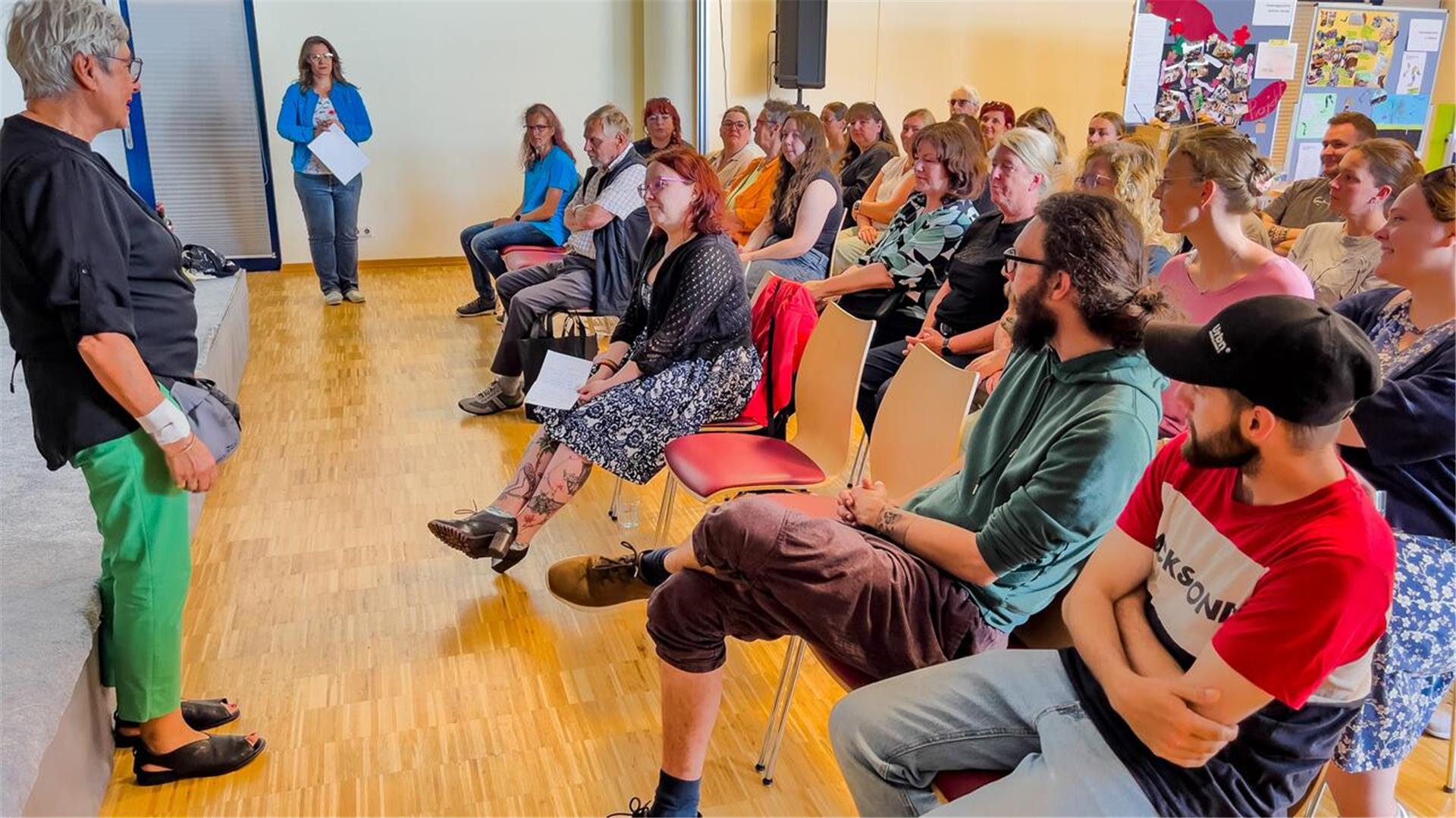 Eine Gruppe von Erwachsenen sitzt in einem Veranstaltungsraum und hört einer Frau im Vordergrund aufmerksam zu. Die Frau steht seitlich zur Kamera und spricht zu den Teilnehmenden. Im Hintergrund sind weitere Personen sowie Plakate und Infowände zu sehen.