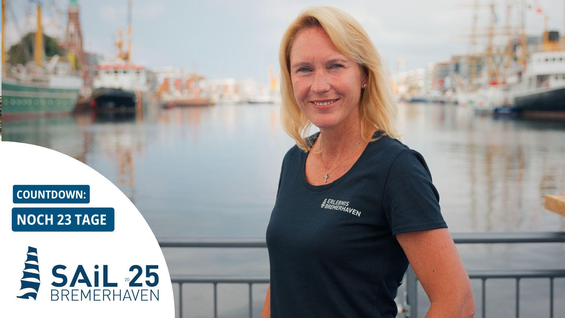 Frau im T-Shirt mit „Erlebnis Bremerhaven“-Logo steht lächelnd am Hafen; im Hintergrund liegen historische Segelschiffe. Links im Bild Countdown zur Sail 2025 Bremerhaven: Noch 23 Tage.