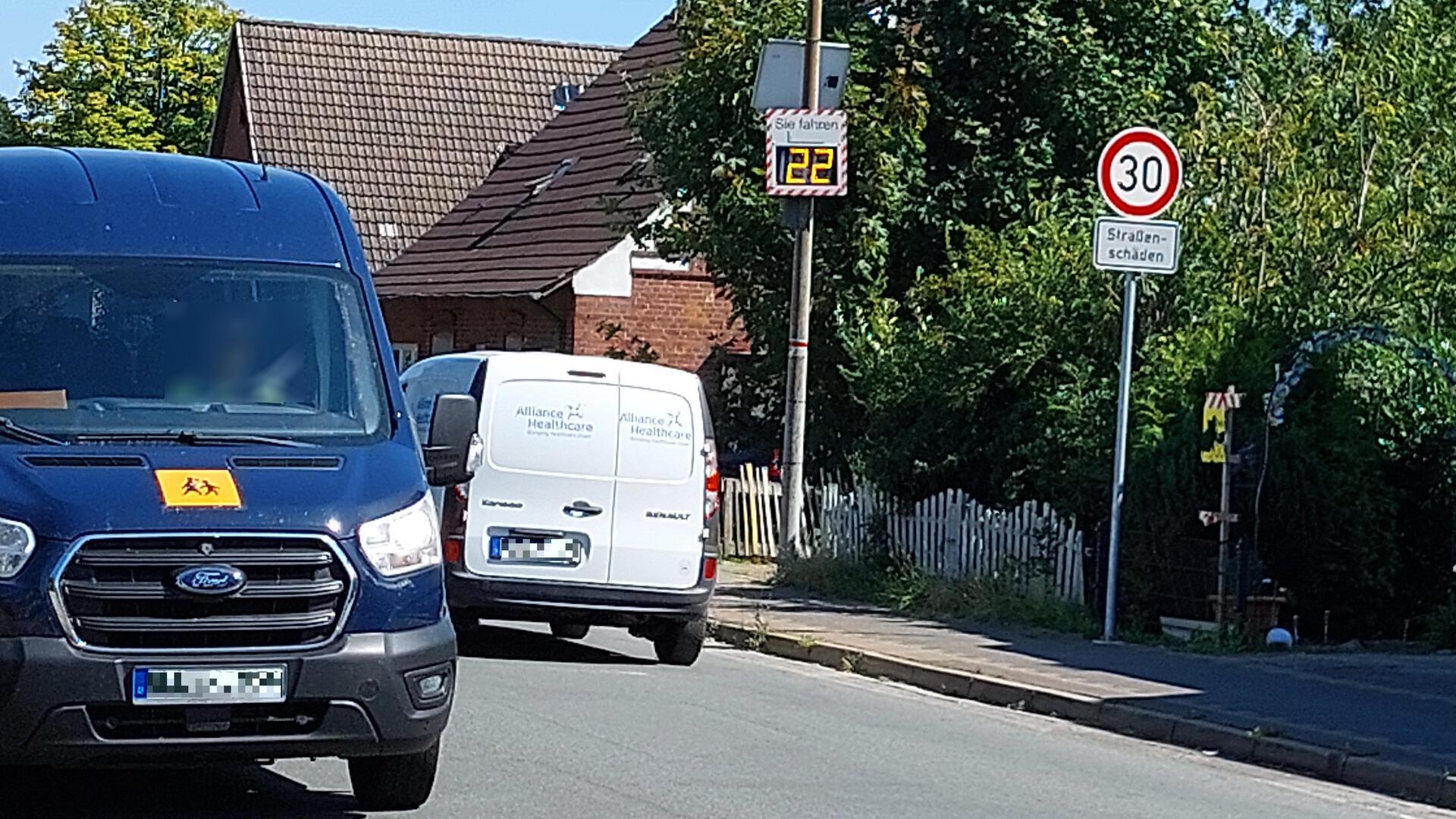 Zwei Lieferwagen fahren auf einer Straße mit Tempo-30-Zone in einem Wohngebiet. Rechts steht ein Verkehrsschild mit der Aufschrift „30 – Straßenschäden“ sowie eine digitale Geschwindigkeitsanzeige, die „22“ km/h zeigt.