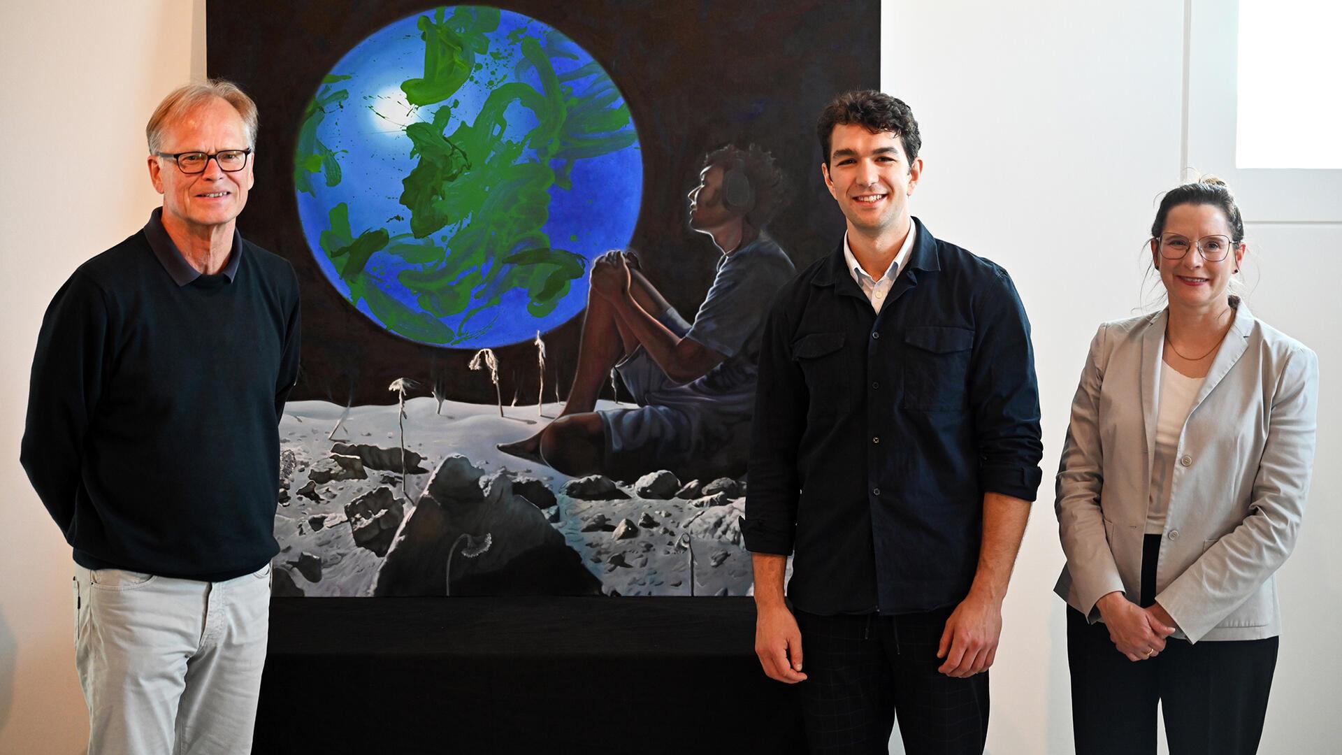 Gruppenfoto mit zwei Herren, einer Frau und einem Gemälde, das sich zwischen den beiden Herren befindet. „The End of Nowhere“ von Paris Giachoustidis (Mitte) ist das erste Kunstwerk der Sonderausstellung „Verlockung Weltall. Auswandern auf Mars, Mond und Venus?“ ist im Deutschen Auswandererhaus. Das Foto zeigt zudem Jörg Schulz und Dr. Simone Blaschka.