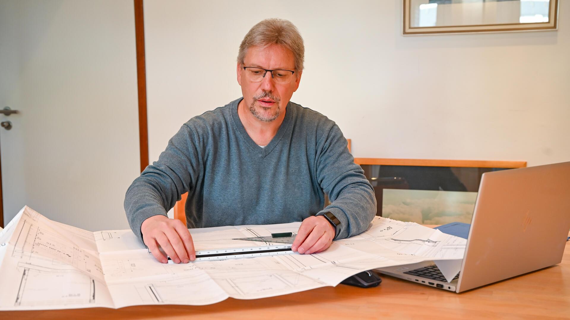 Ein Mann sitzt an einem Schreibtisch, auf dem Papier, Laptop und Lineale liegen. 