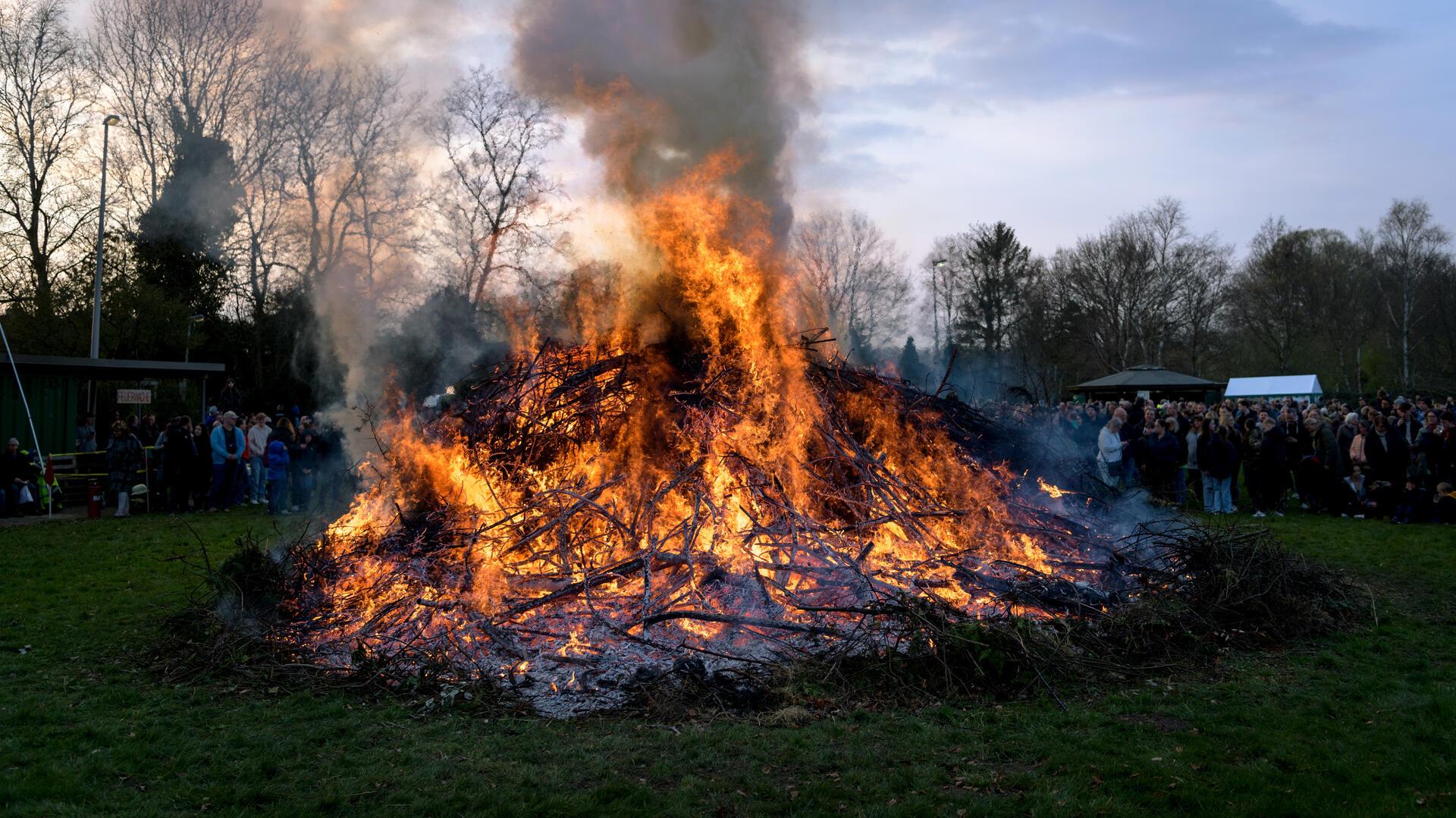 Großes Osterfeuer aus brennenden Ästen und Zweigen auf einer Wiese, umgeben von zahlreichen Zuschauerinnen und Zuschauern im Hintergrund; dichter Rauch steigt in den abendlichen Himmel, während die Flammen hell lodern.