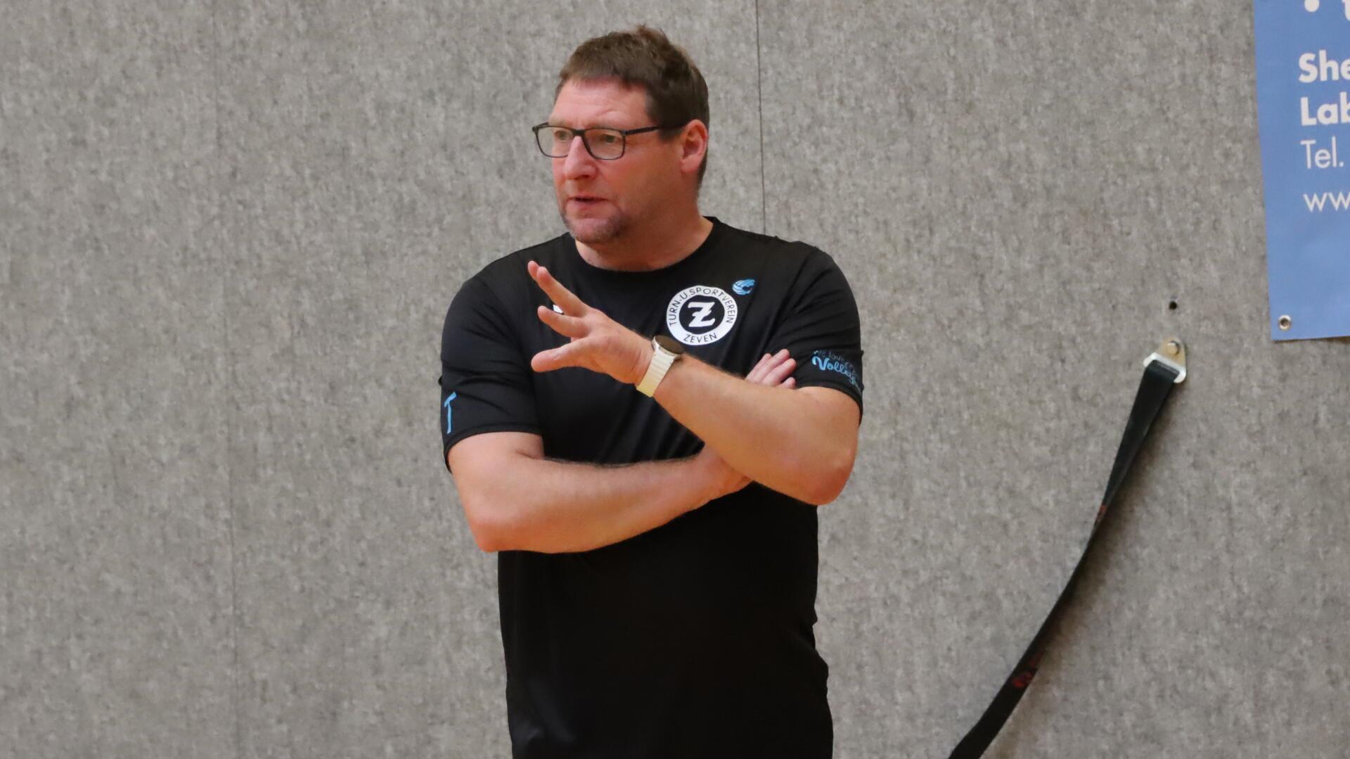 Ein Mann schaut mit verschränkten Armen gespannt einem Volleyballspiel zu. 