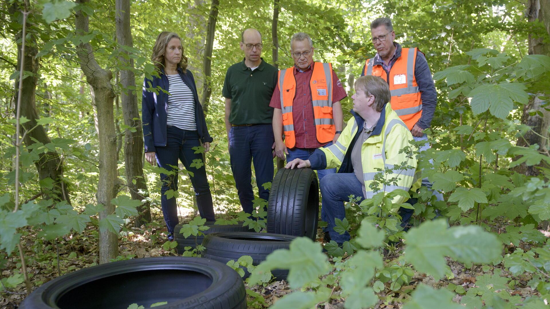 Fünf Personen, darunter eine Frau und vier Männer, stehen und knien im Wald neben illegal entsorgten Autoreifen. Drei der Männer tragen Warnkleidung.