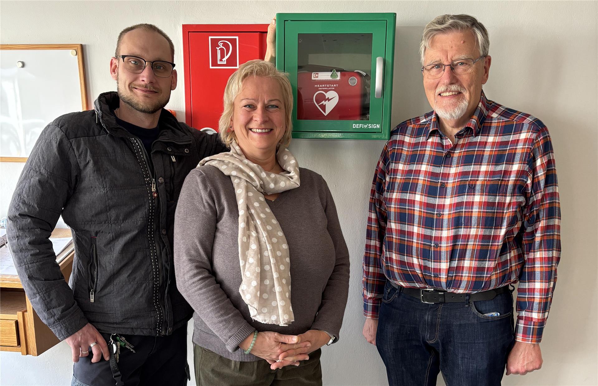 Drei Personen stehen lächelnd vor einer weißen Wand mit einem grünen Defibrillatorkasten und einem roten Feuerlöscherkasten.