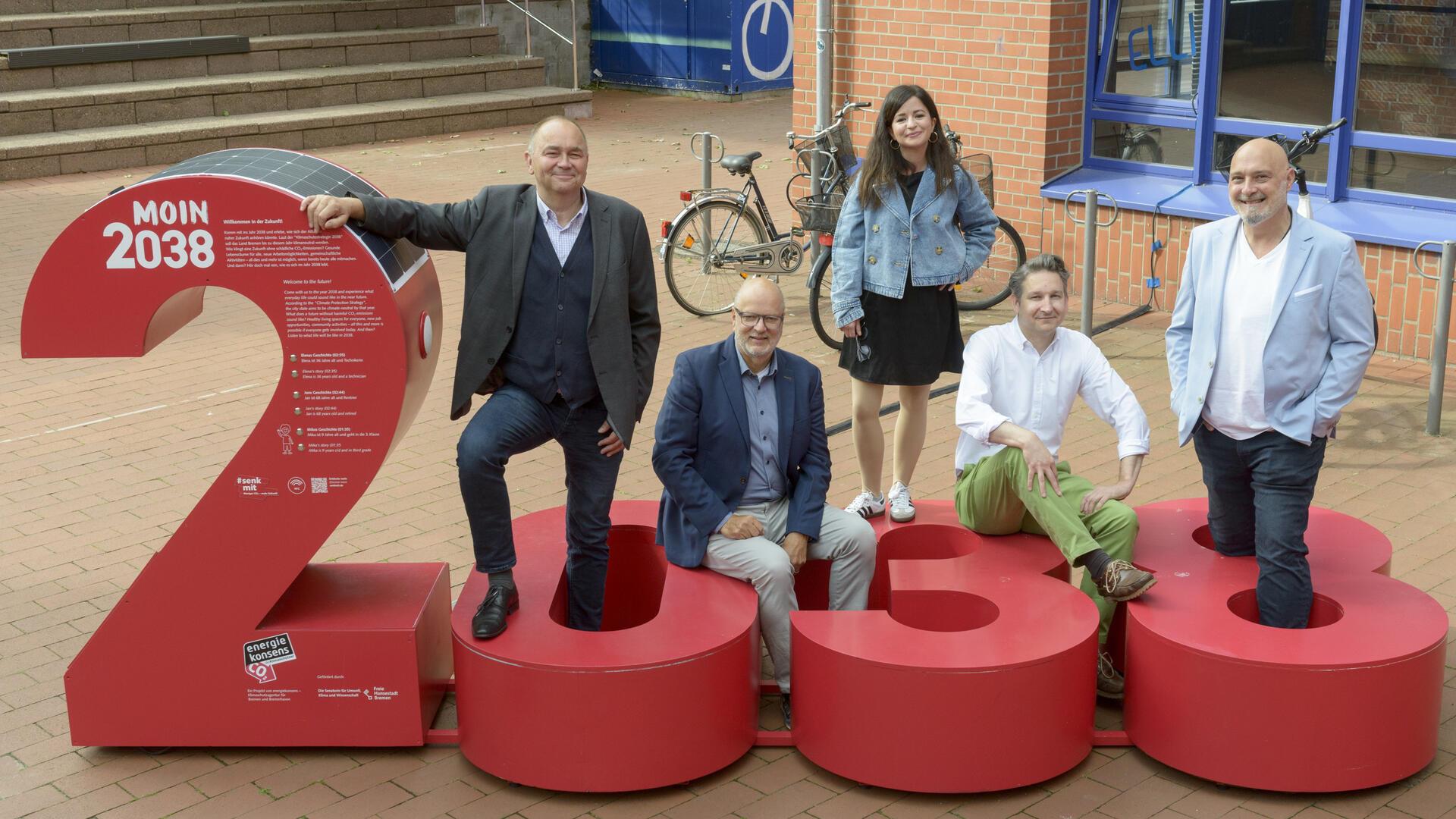 Fünf Personen stehen und sitzen um die rote Installation „MOIN 2038“ auf dem Campus der Hochschule Bremerhaven, die symbolisch für das Klimaziel des Landes Bremen steht.