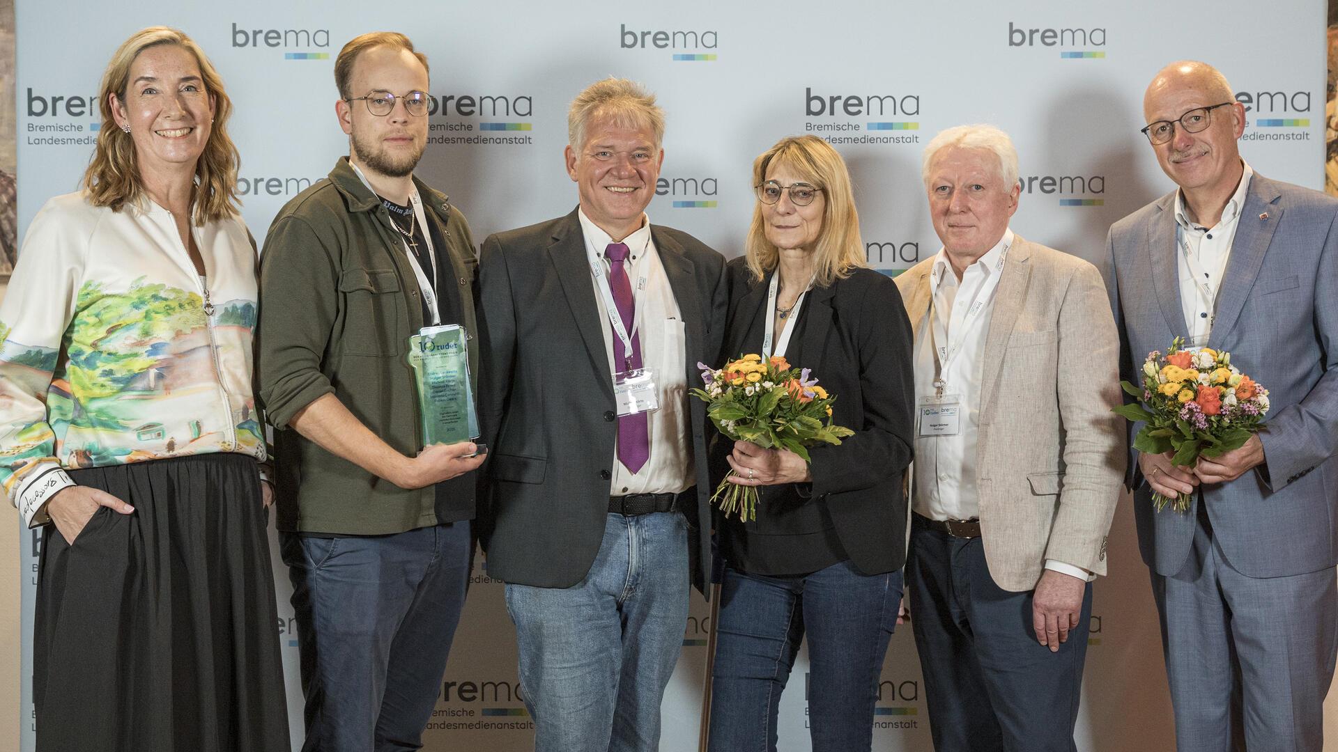 Gruppenfoto bei der Preisverleihung der Bremischen Landesmedienanstalt: Sechs Personen stehen vor einer Fotowand, drei von ihnen halten Blumensträuße oder eine Trophäe in den Händen und lächeln in die Kamera.