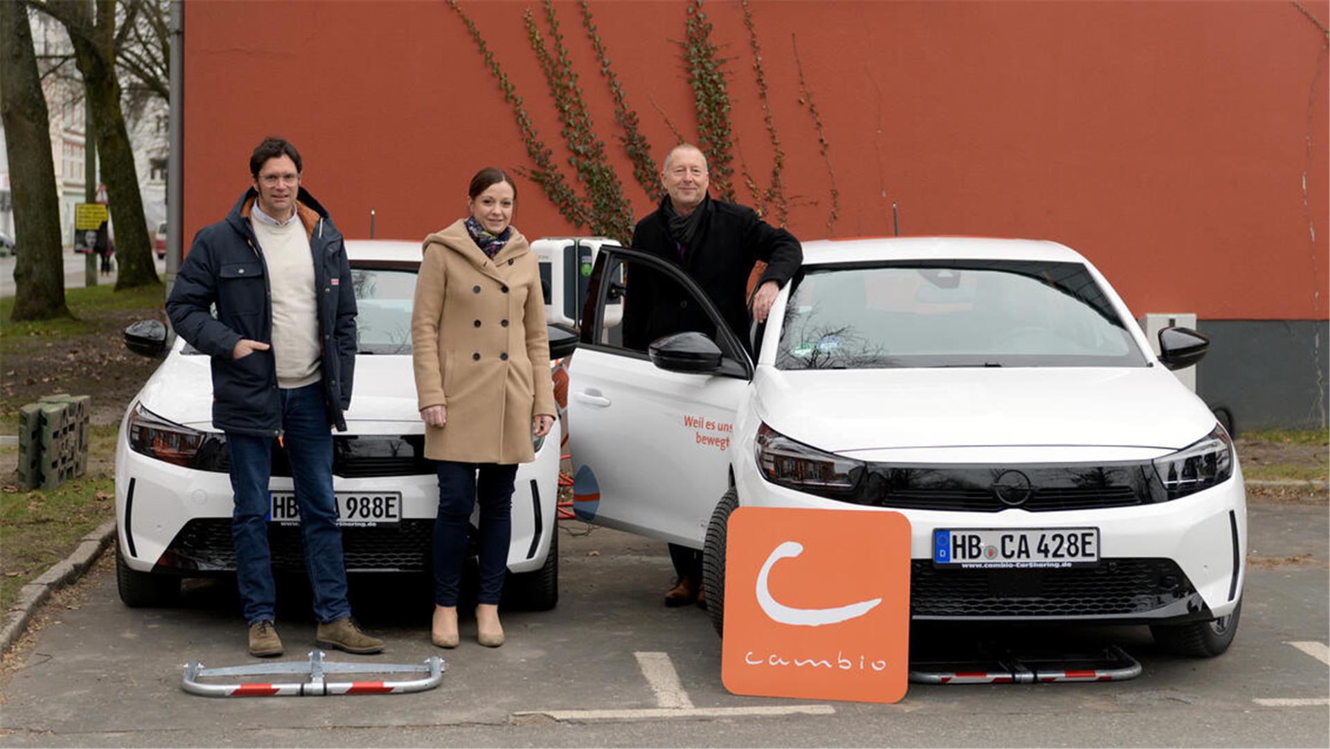 Drei Personen stehen vor zwei weißen Elektroautos von cambio CarSharing. Eine Person lehnt an der offenen Autotür, während die anderen daneben posieren. Ein orangefarbenes cambio-Logo steht vor dem Fahrzeug. Im Hintergrund eine rote Wand mit Efeu.