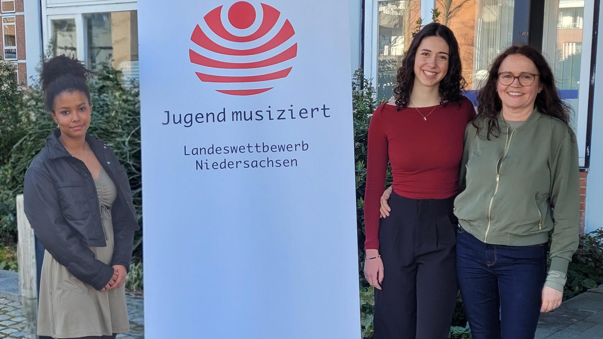 Zwei junge Frauen und eine Erwachsene stehen vor einem Gebäude neben einem Banner mit der Aufschrift „Jugend musiziert – Landeswettbewerb Niedersachsen“.