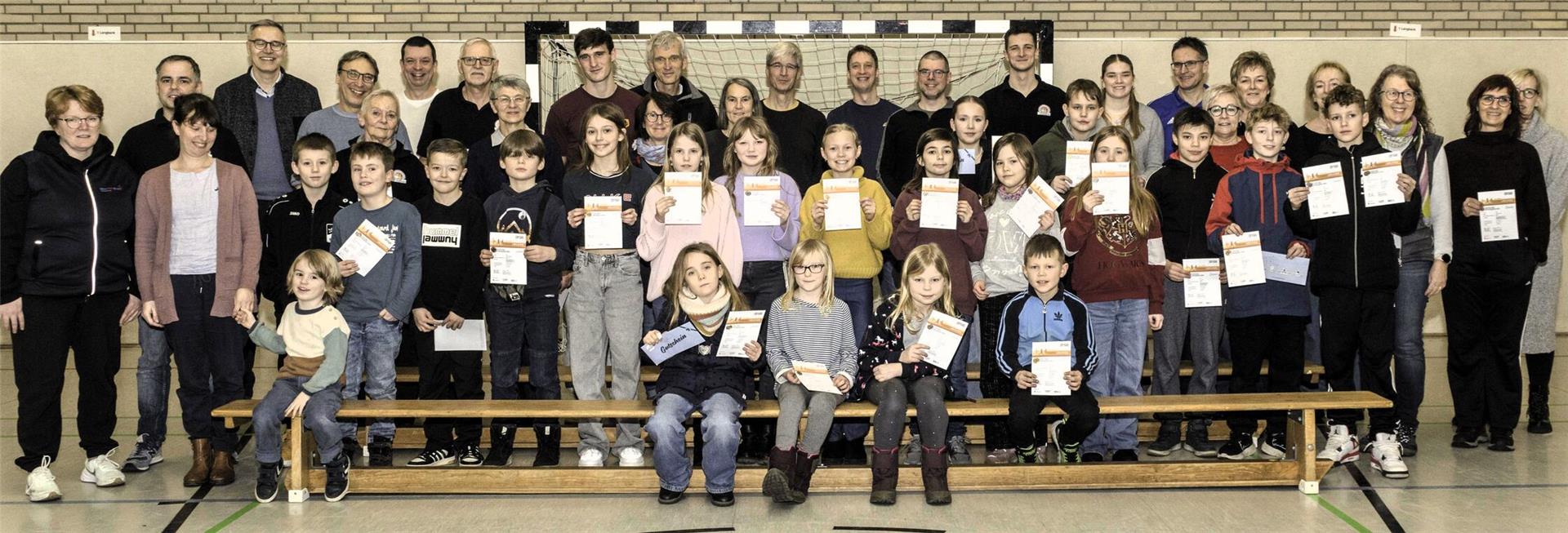 Eine große Gruppe von Menschen, bestehend aus Erwachsenen und Kindern, posiert in einer Turnhalle für ein Gruppenfoto. Die Kinder in der vorderen Reihe halten Urkunden in den Händen, während die Erwachsenen dahinter stehen. Im Hintergrund ist ein Handballtor und eine helle Wand aus Klinkersteinen sichtbar.