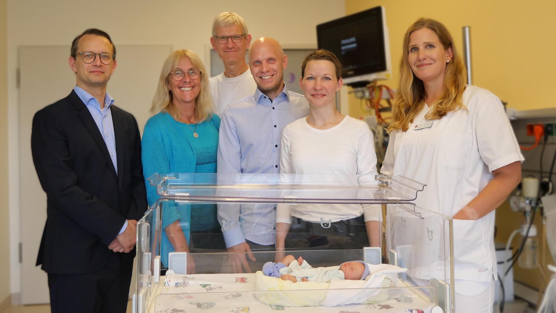 Sechs Erwachsene, darunter medizinisches Personal und weitere Personen, stehen lächelnd in einem Krankenhauszimmer hinter einem Inkubator mit der Puppe eines neugeborenen Babys darin.