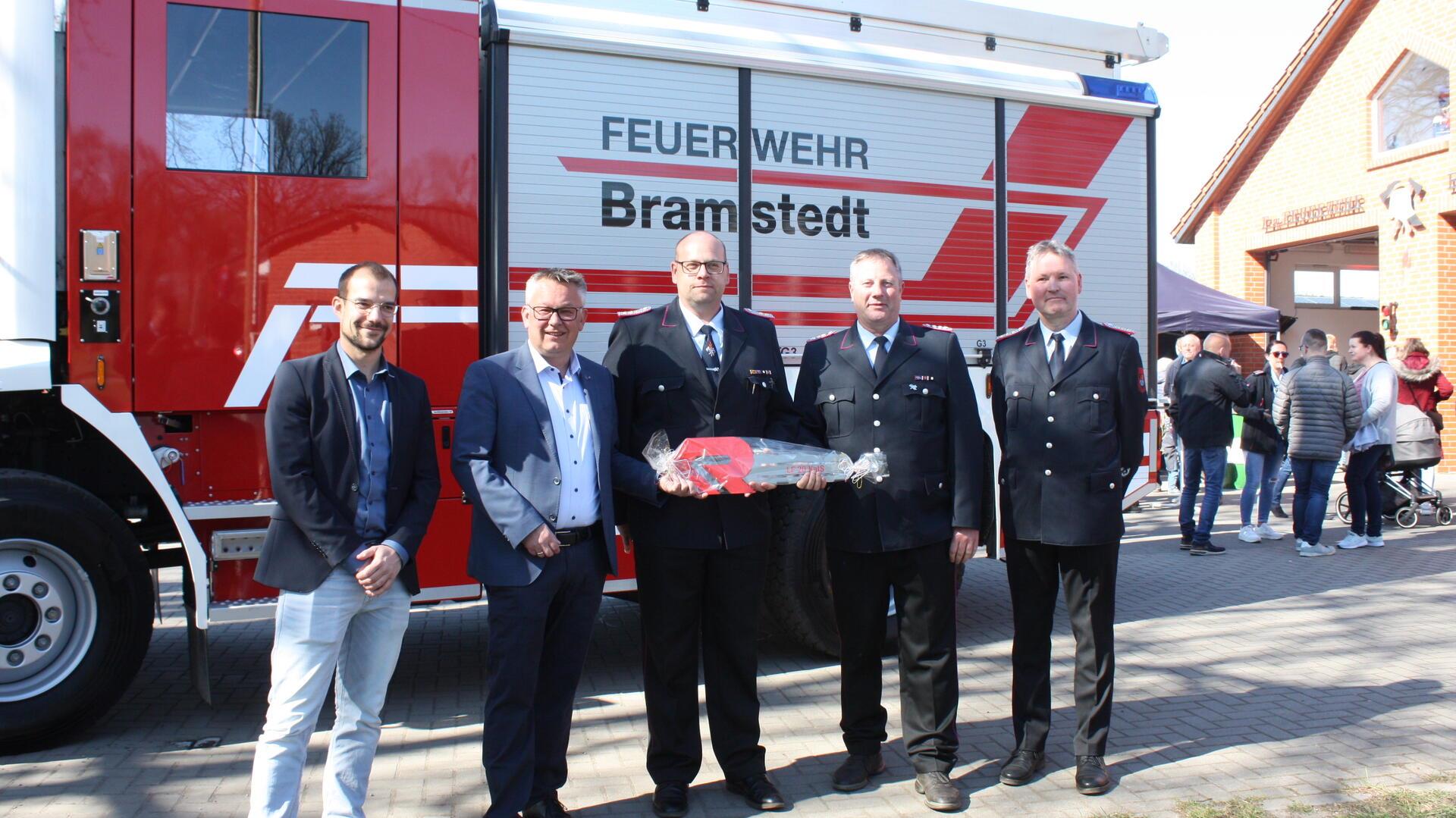 Fünf Männer, darunter Feuerwehrleute in Uniform und Vertreter der Gemeinde, stehen vor einem neuen Feuerwehrfahrzeug mit der Aufschrift „Feuerwehr Bramstedt“. Einer der Feuerwehrmänner hält symbolisch einen großen, verpackten Fahrzeugschlüssel. Im Hintergrund sind Besucherinnen und Besucher beim Tag der offenen Tür an der Fahrzeughalle zu sehen.