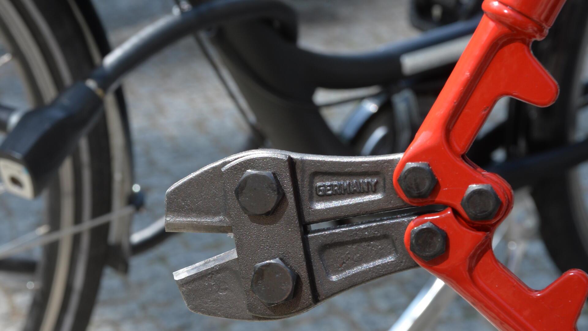 Ein Bolzenschneider wird vor ein mit einem Fahrradschloss gesichertes Fahrrad gehalten.
