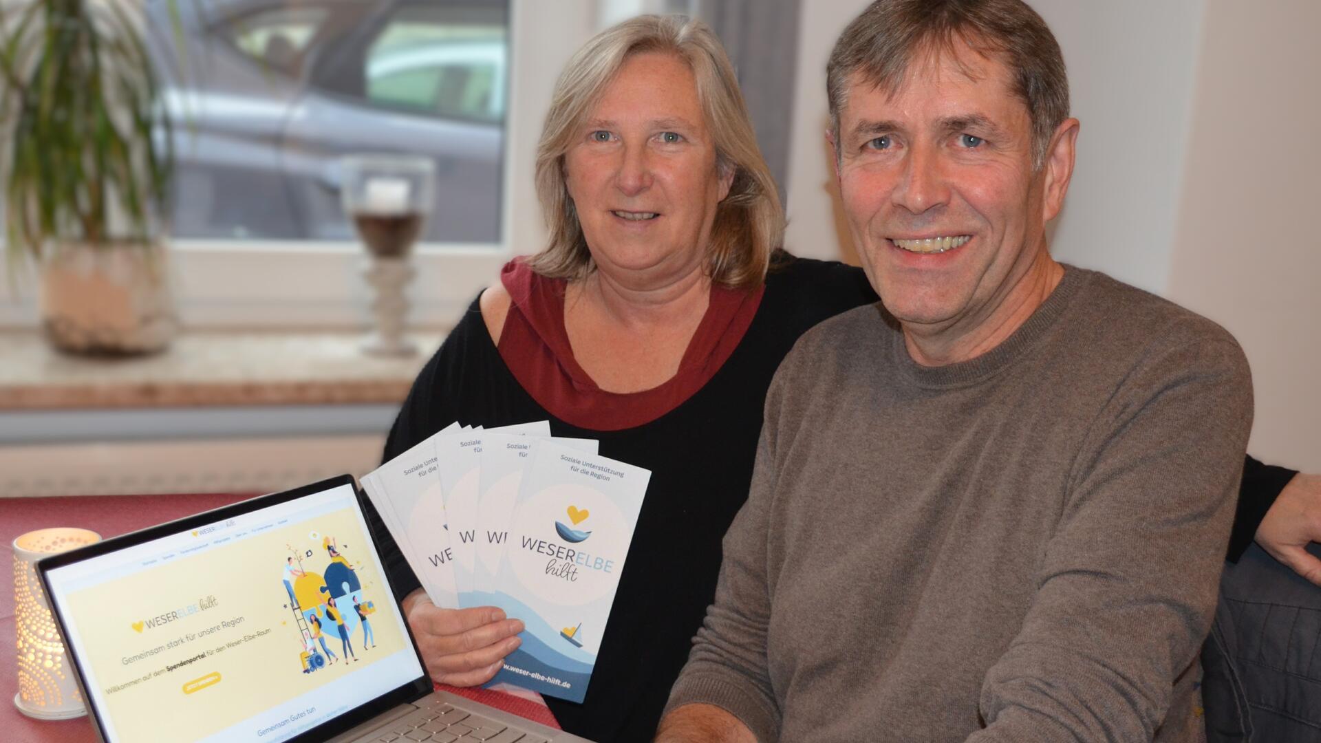 Eine Frau und ein Mann sitzen an einem Tisch. Die Frau hält Flyer von "Weser-Elbe hilft" in der Hand, auf dem Tisch steht ein Laptop, auf dem die Webseite der Organisation geöffnet sit.