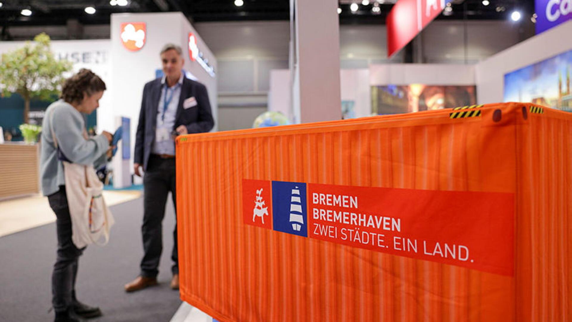 Ein Mann und eine Frau stehen neben einem Mini-Container an einem Messestand.