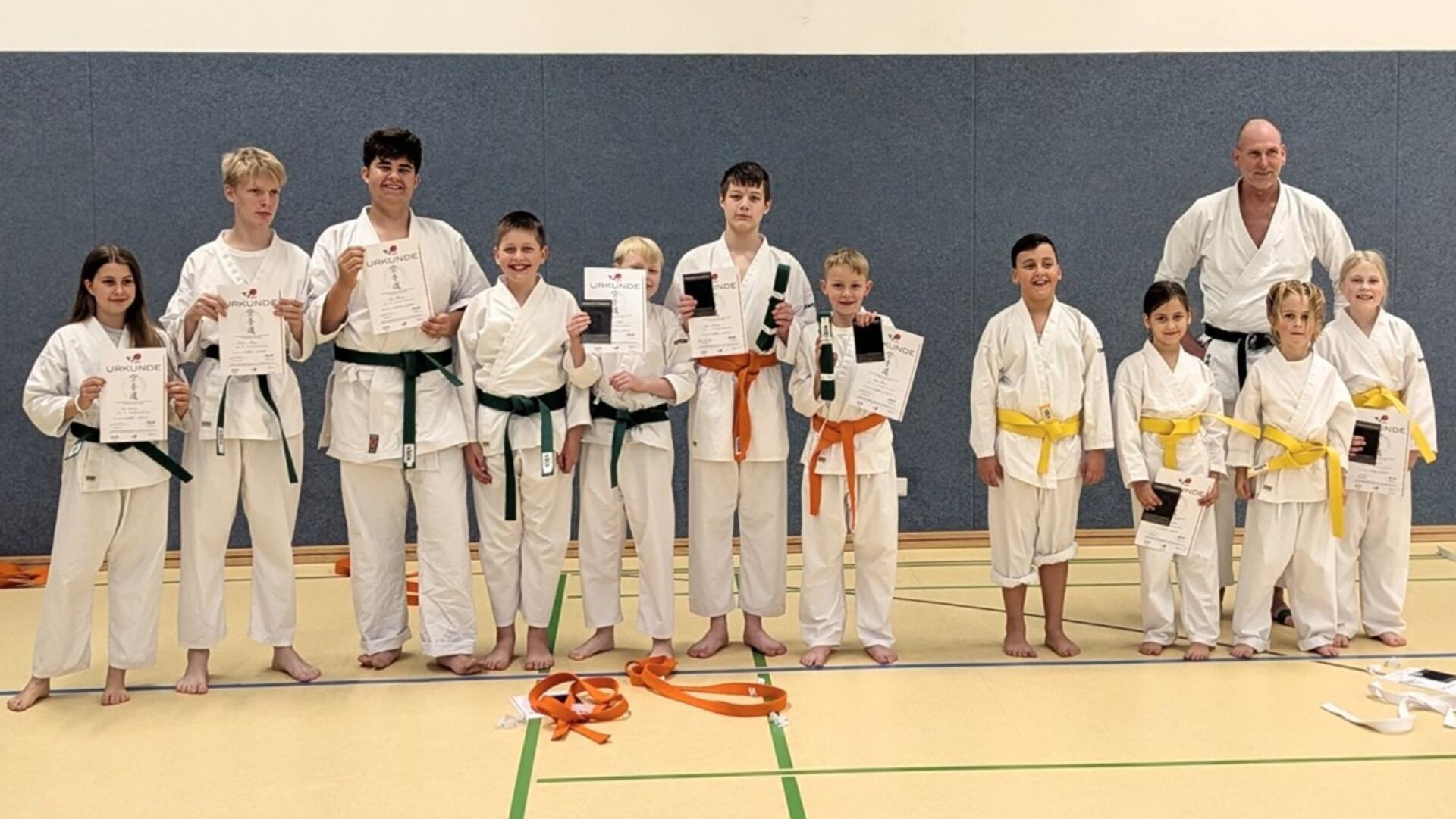 Eine Gruppe von Karatekindern steht in einer Turnhalle nebeneinander und hält Urkunden in den Händen. Einige tragen gelbe, orange oder grüne Gürtel, rechts steht ihr Trainer.
